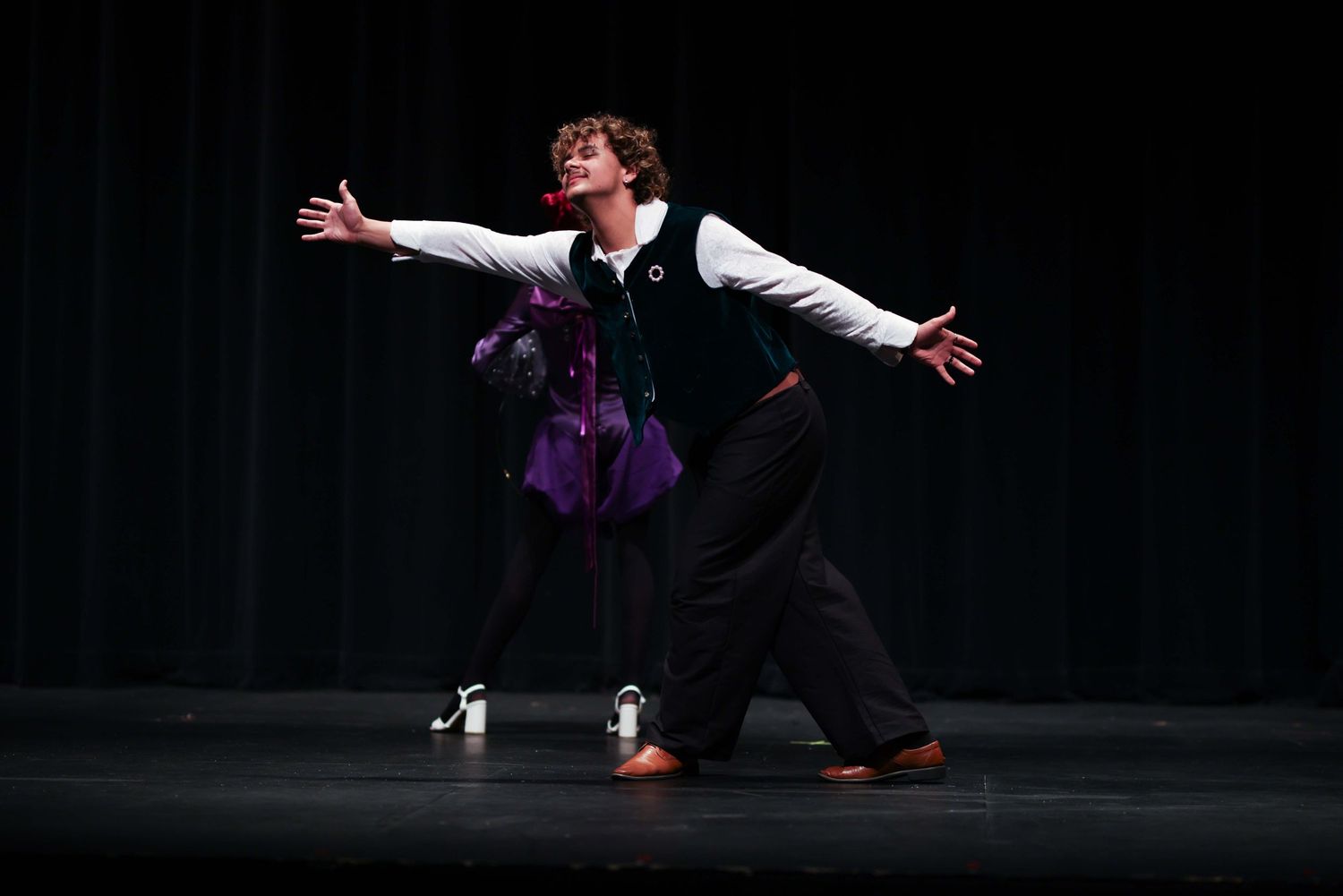 A figure performs a dramatic dance movement in a dark theater with arms outstretched against a black background.