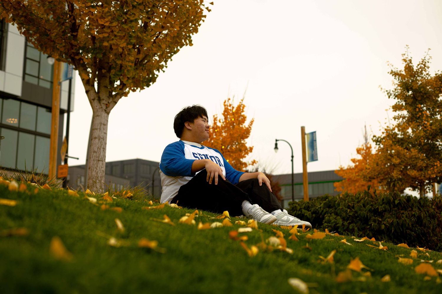 Person relaxes on autumn hillside with fallen leaves and urban buildings visible in distance.