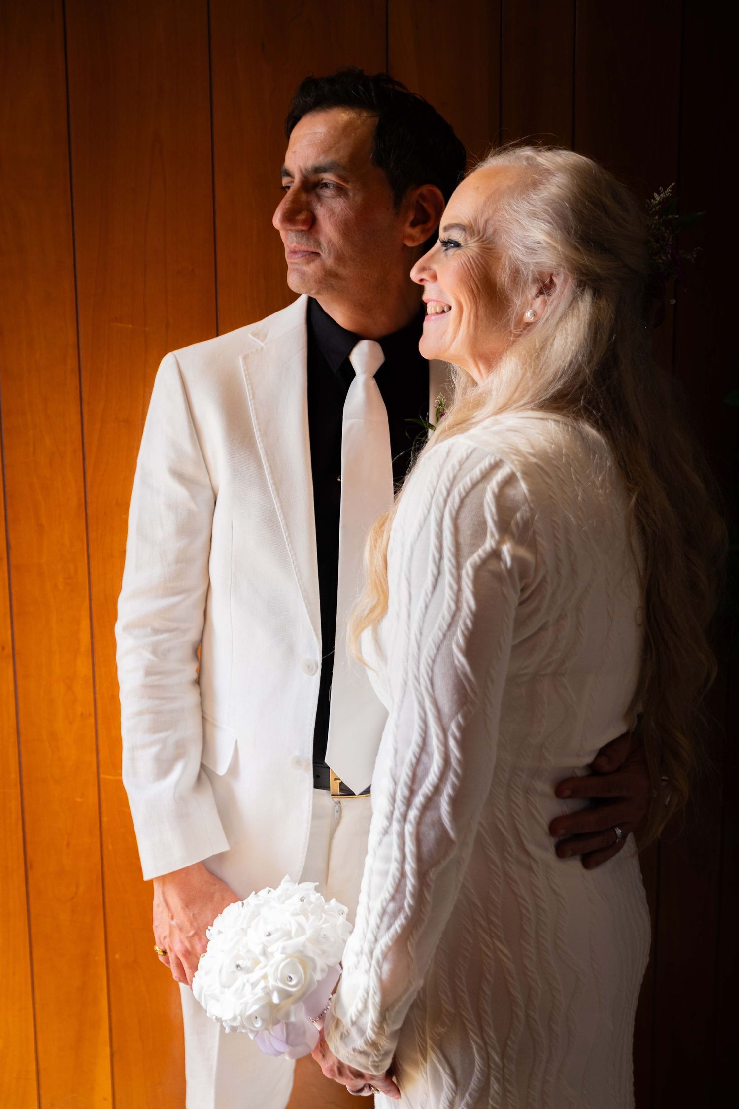 A couple in matching white outfits share an intimate moment against a wooden wall with warm lighting.