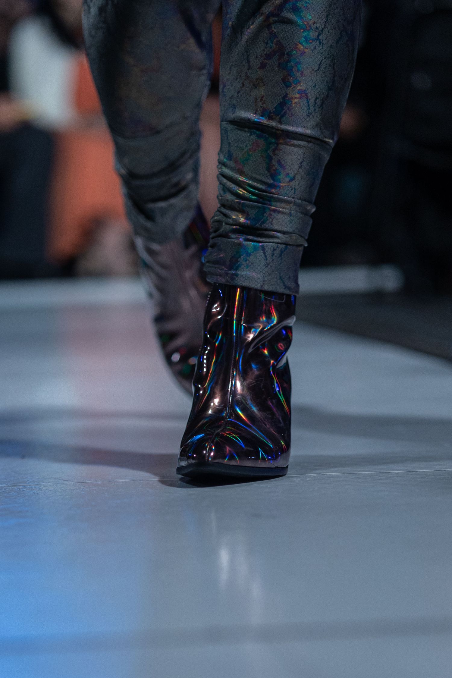 Close-up of shiny black leather boots walking on a fashion runway with blue lighting.