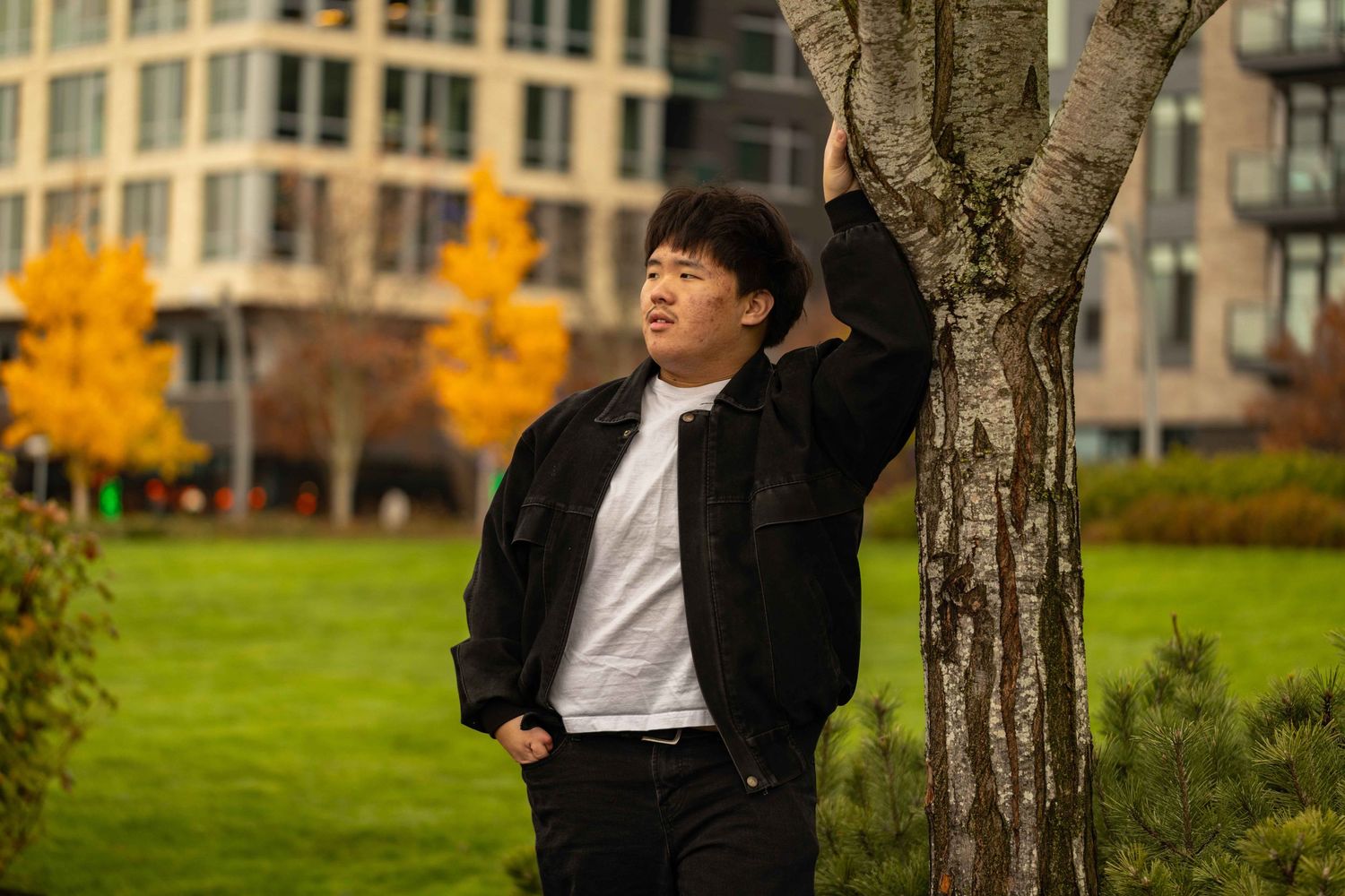 Person in black suit jacket sits on grassy hill during autumn with golden trees and buildings in background.