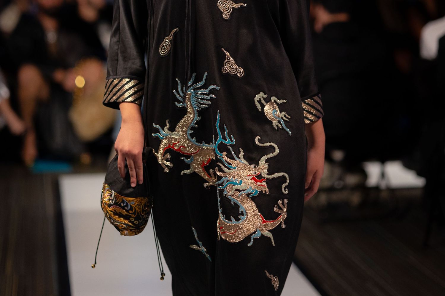 A fashion model walks down the runway wearing a black dress with dragon embroidery at a fashion show.