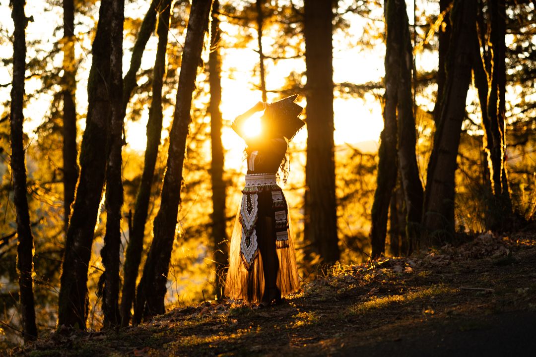 A woman in a flowing boho dress stands among tall trees bathed in golden sunset light in a forest.