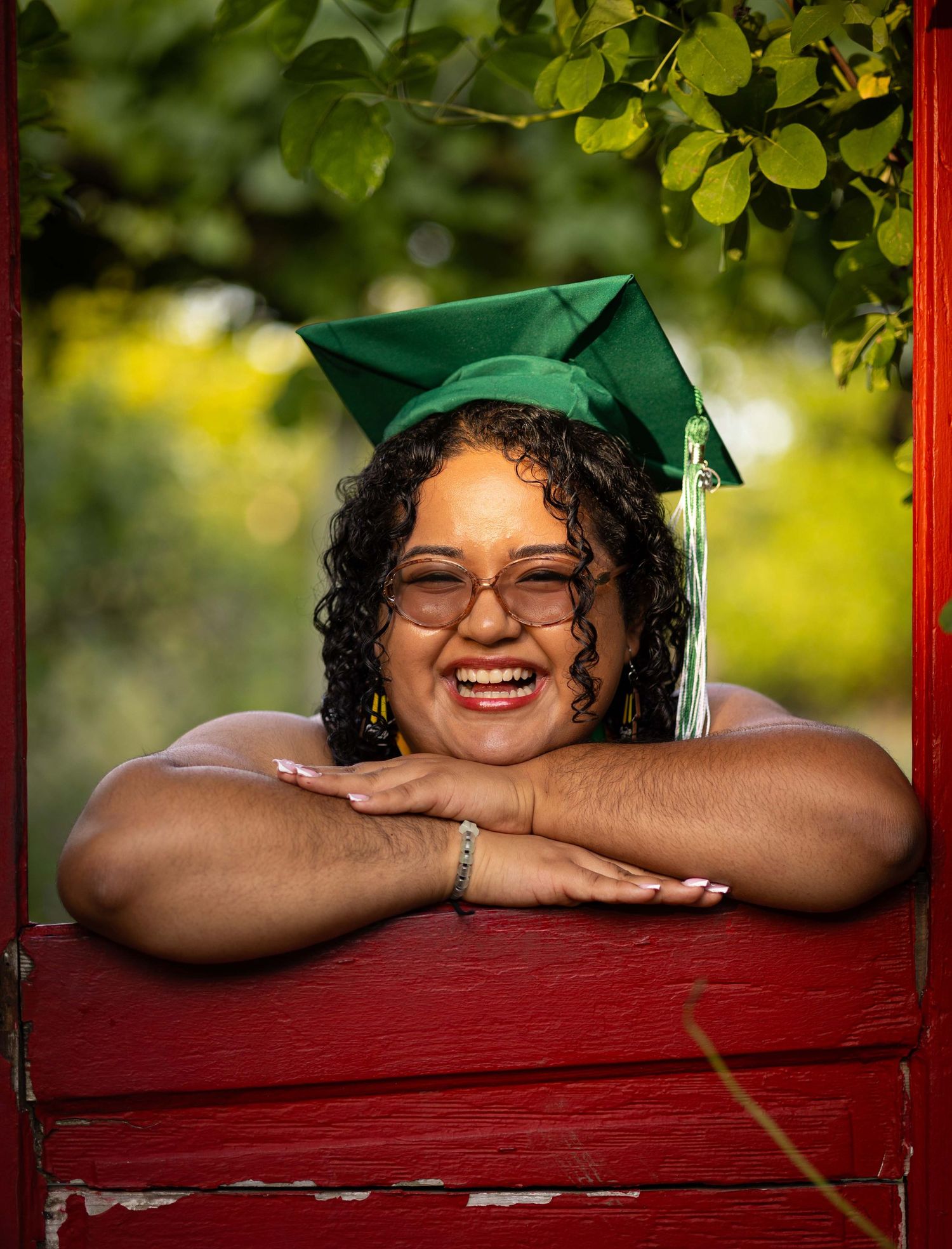 Graduate in cap and gown resting on red bench with natural background.