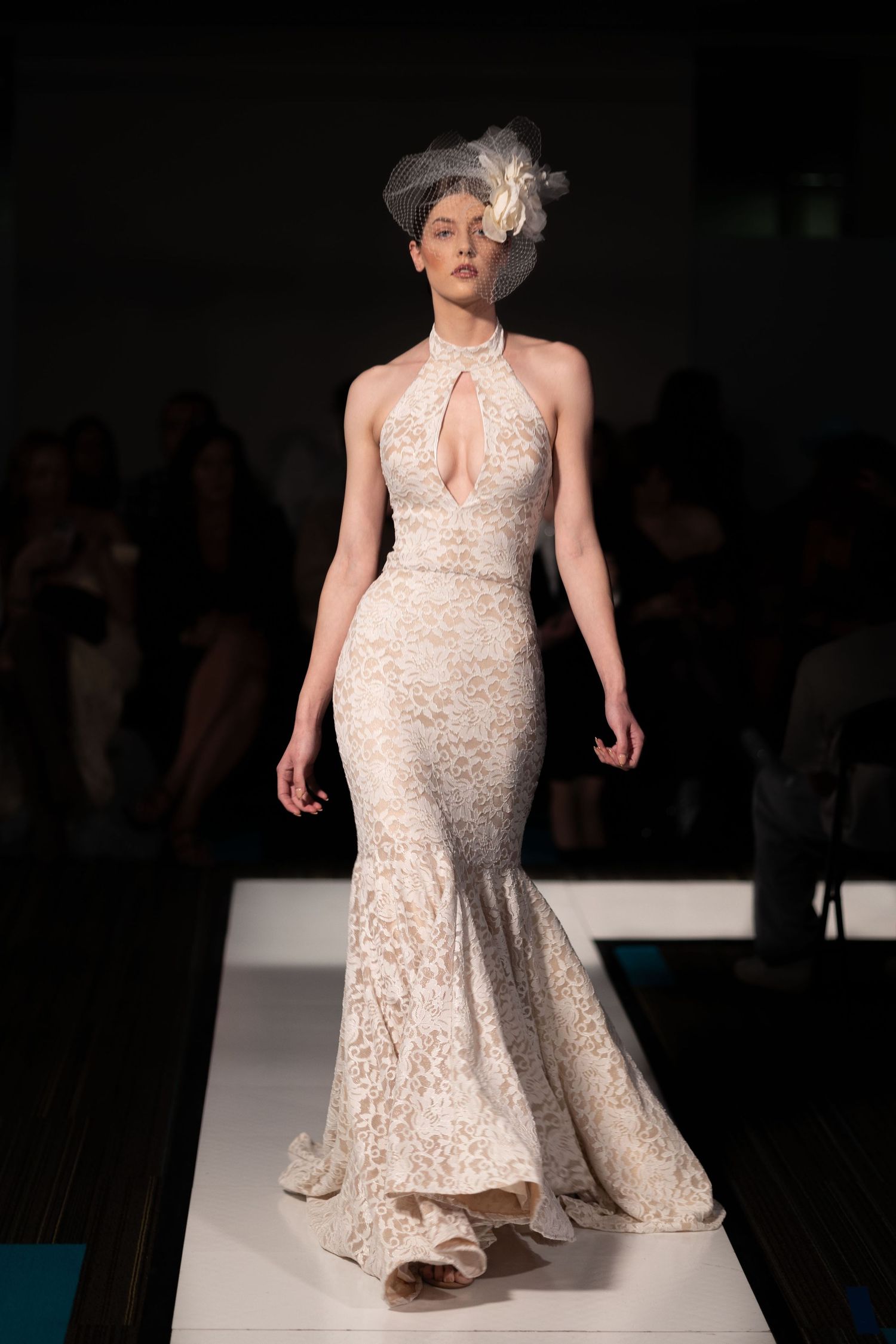 Model in cream lace gown walking down white runway at fashion show.