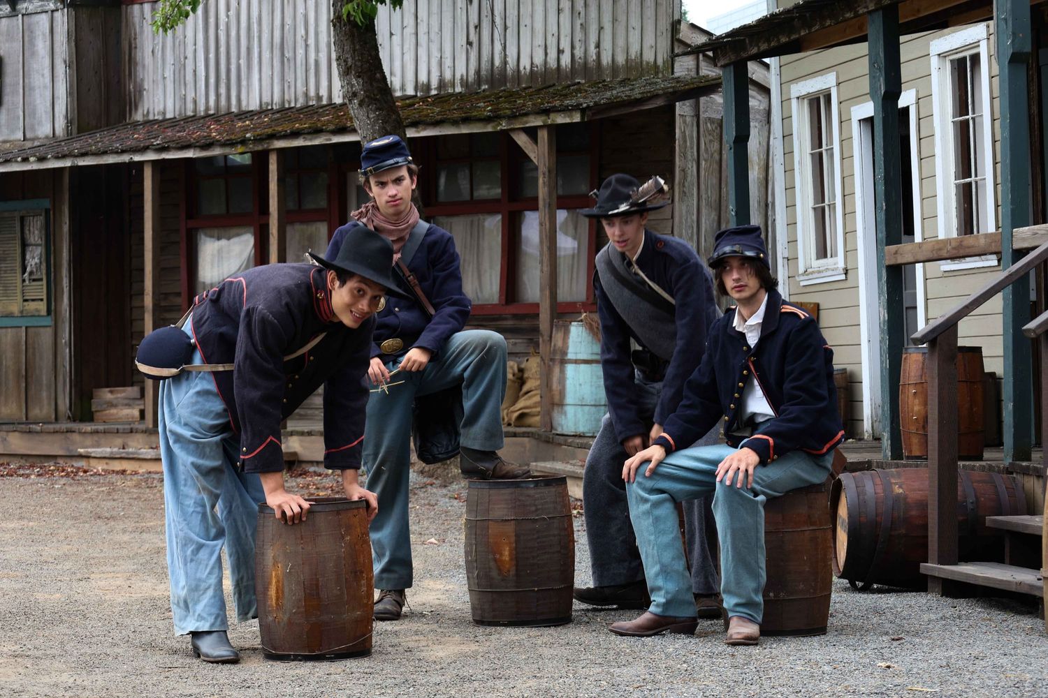 People in denim gather around wooden barrels outside a rustic building with corrugated metal siding.