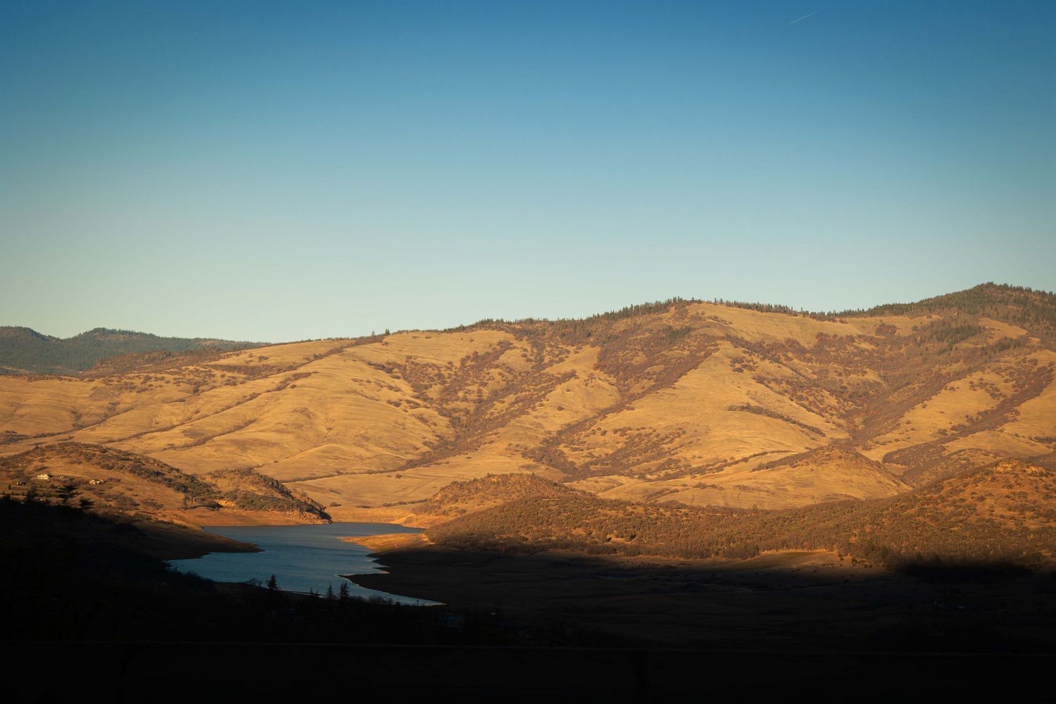 Golden sunlight illuminates rolling hills and mountains against a clear blue sky at sunset.
