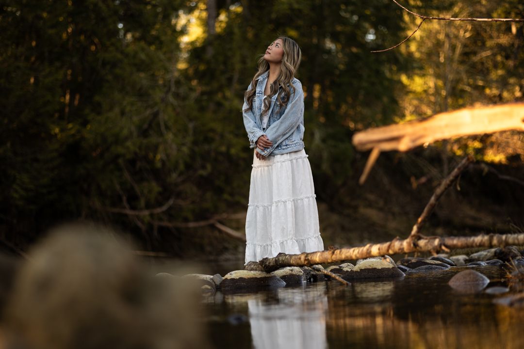 Woman in white skirt and denim jacket standing on rocks by a forest stream, gazing upward in serene nature setting.