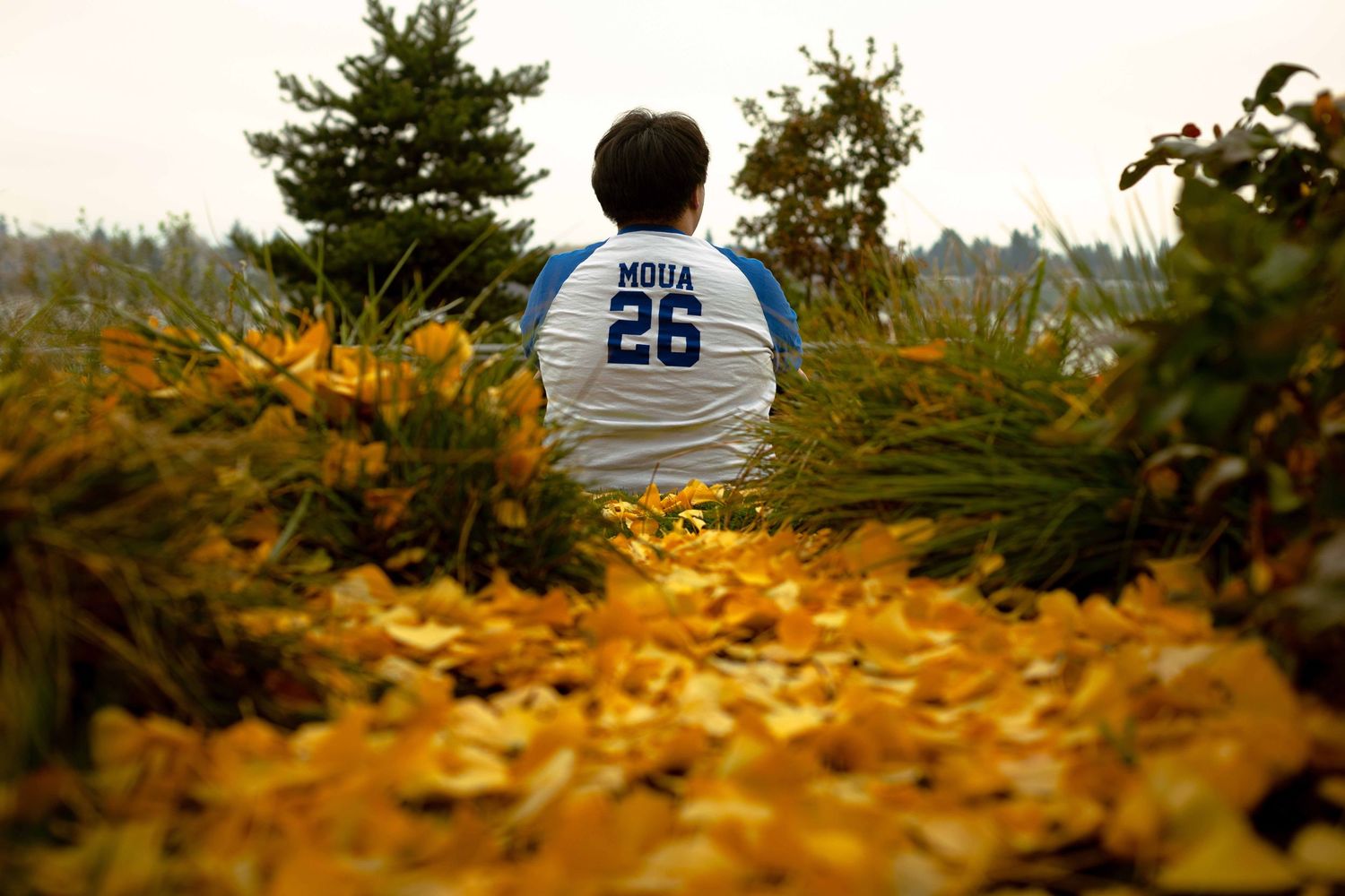 Someone wearing a white jersey sits among fallen autumn leaves on a path surrounded by trees and foliage.