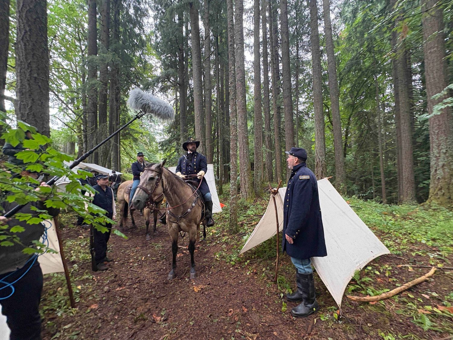 Group of hikers setting up camp with tarps and gear in a dense forest of tall evergreen trees.
