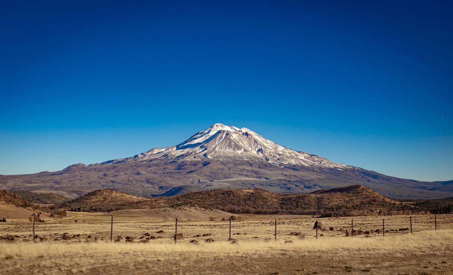 Panoramic view of snow-capped Mount Shasta rising above arid landscape under clear blue skies in California.