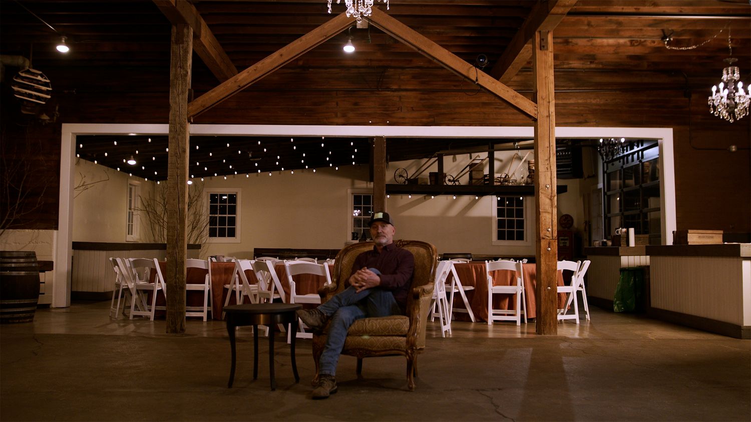 Man sitting alone in armchair inside rustic barn wedding venue with string lights and white folding chairs.