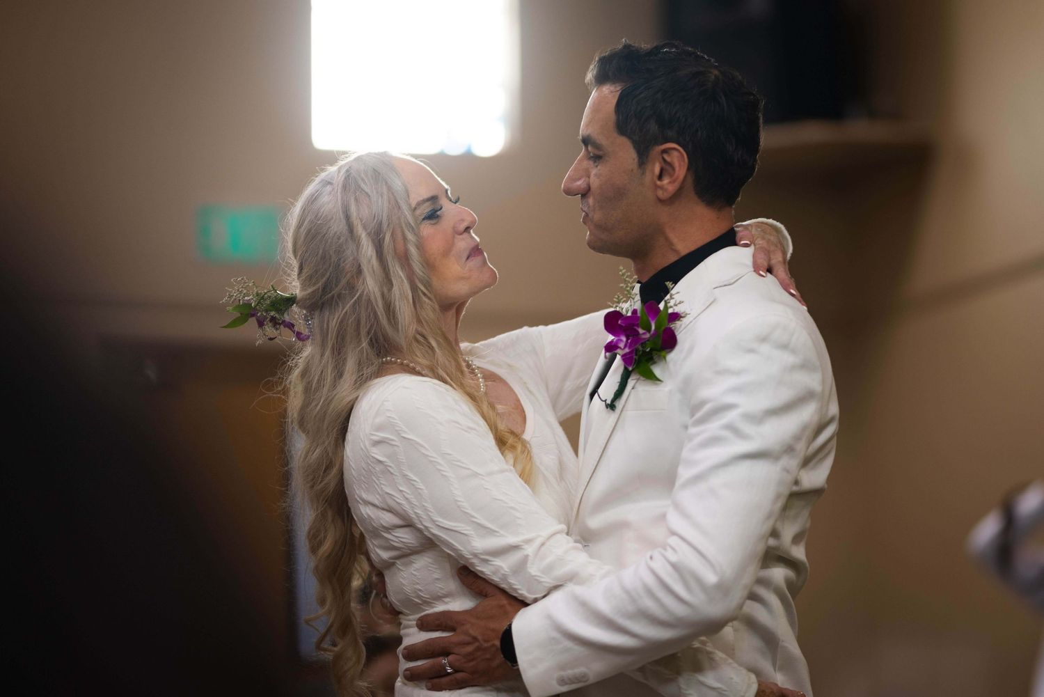 Intimate wedding moment captured in soft lighting as couple embraces in formal white attire.