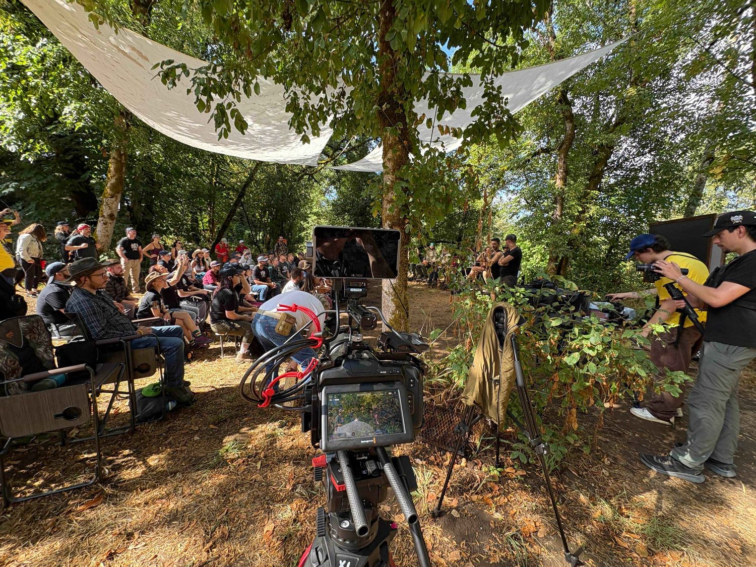 Film crew and equipment set up under shady trees during an outdoor video production shoot.
