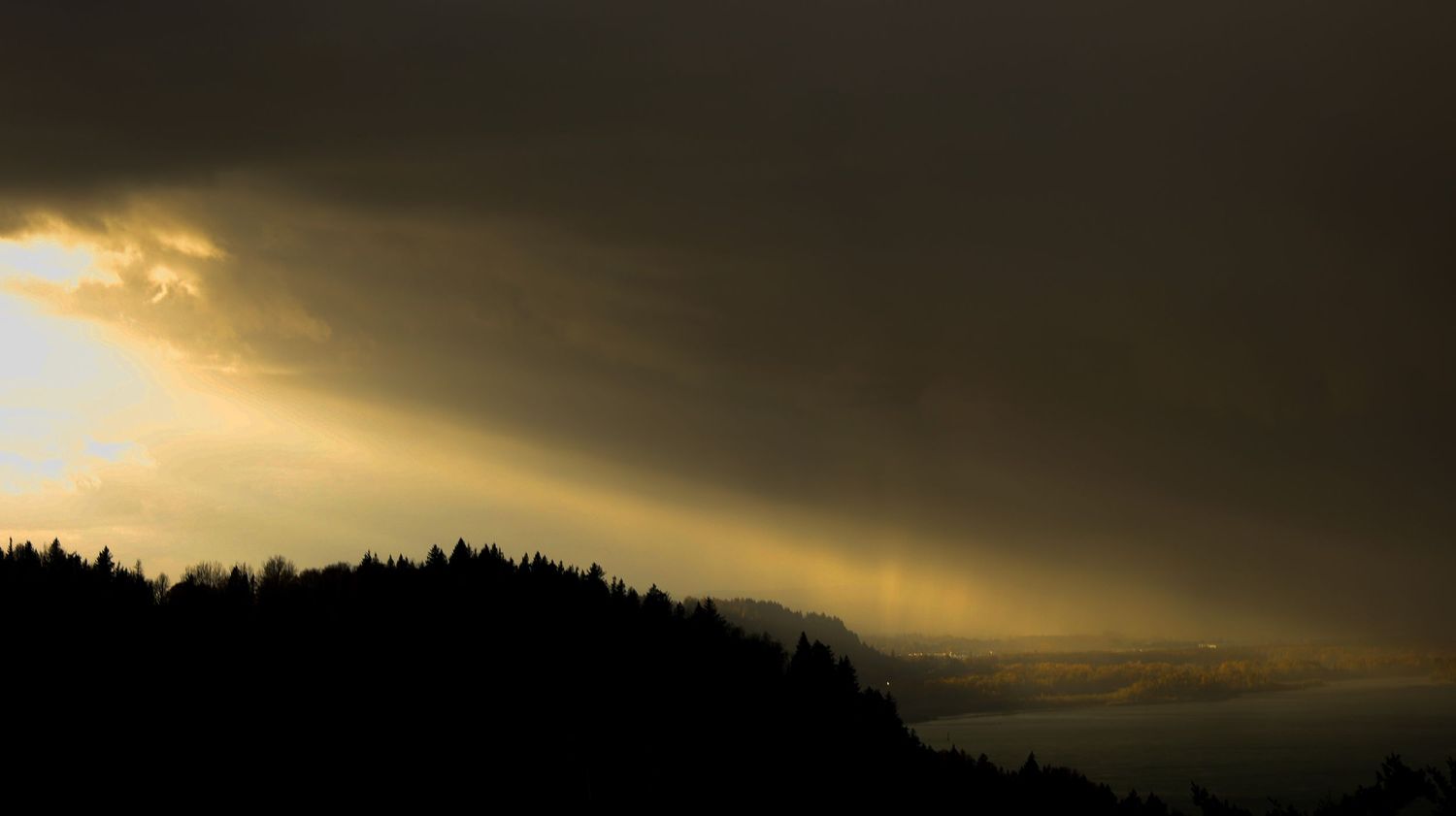 Dramatic sunset sky with sunbeams breaking through dark clouds over a silhouetted forest mountain landscape.