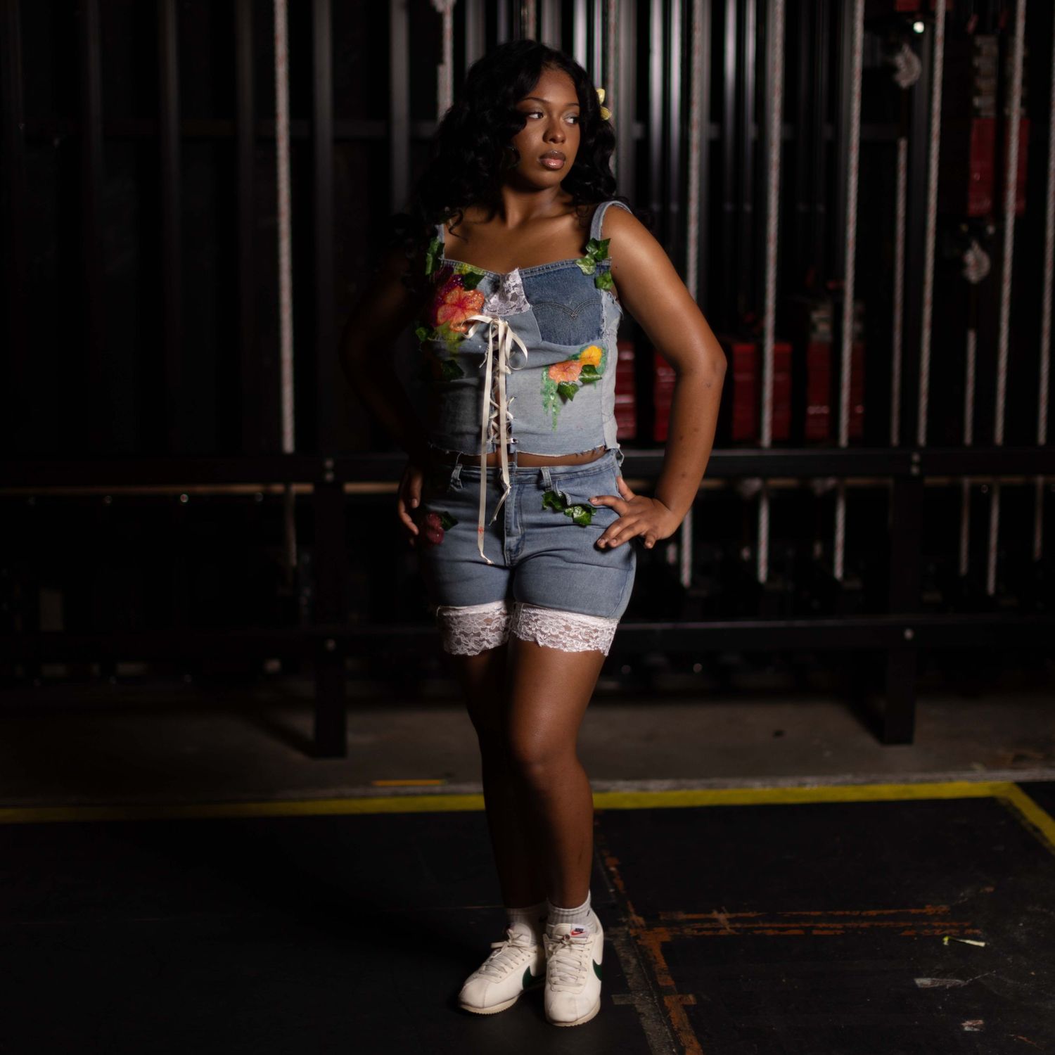 Person in denim shorts and white sneakers poses against dark metal bars in a dimly lit setting.