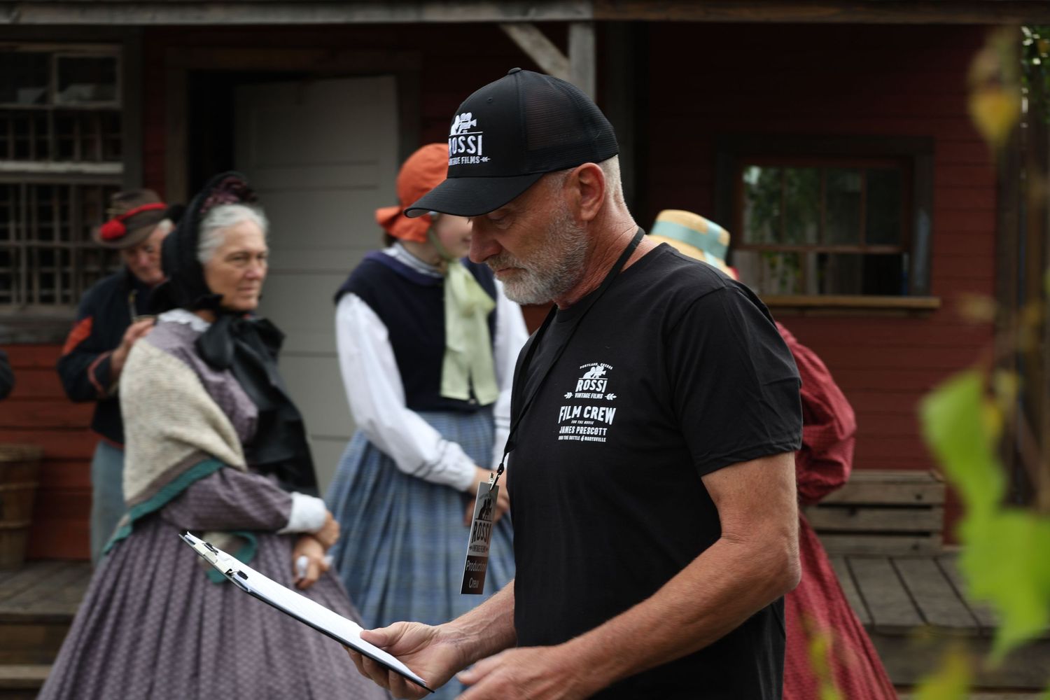 People in period costumes interact during a historical reenactment event at a rustic wooden building.