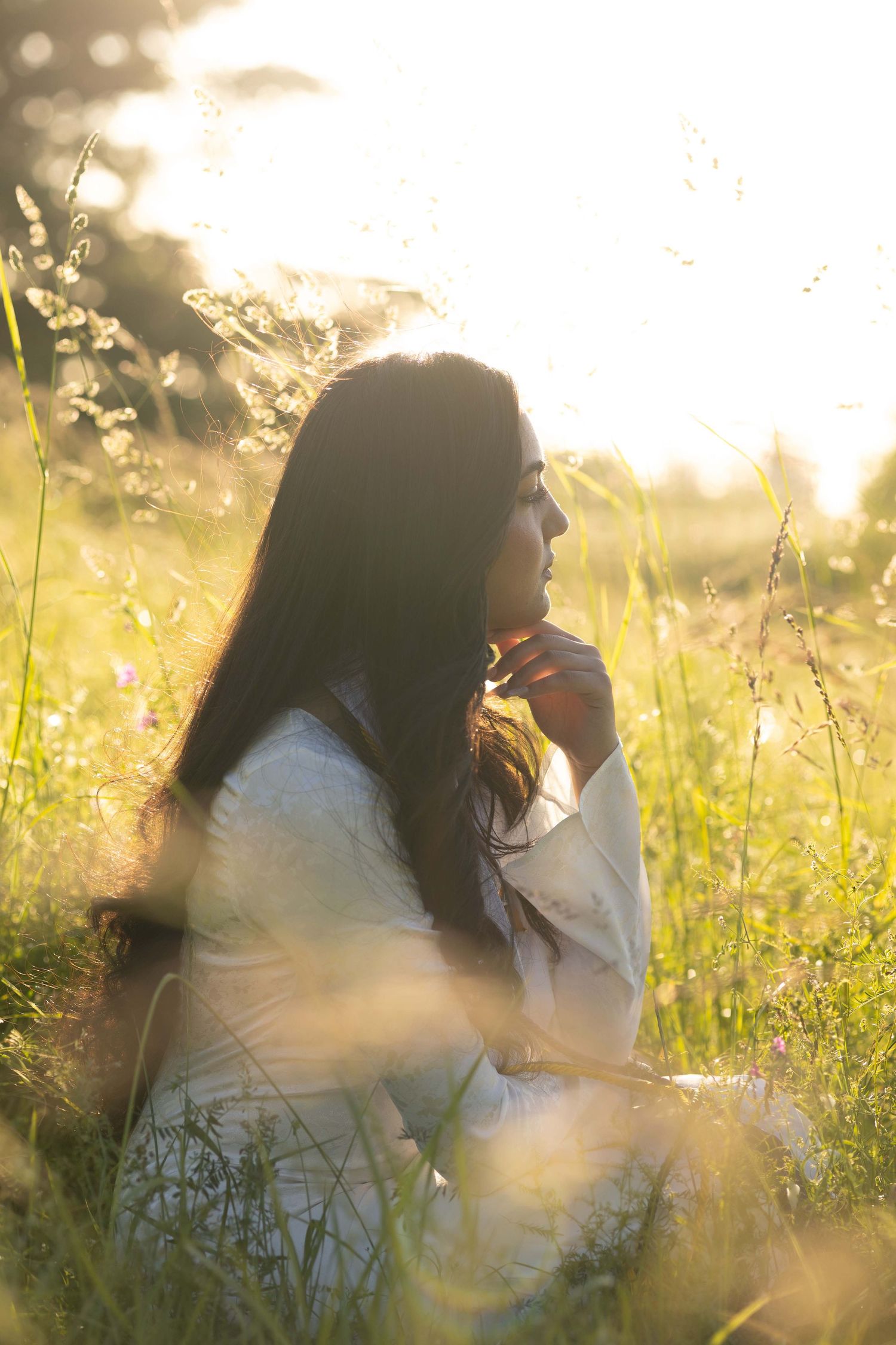 A dreamy golden-hour scene shows a figure sitting in a meadow of tall grass and wildflowers at sunset.