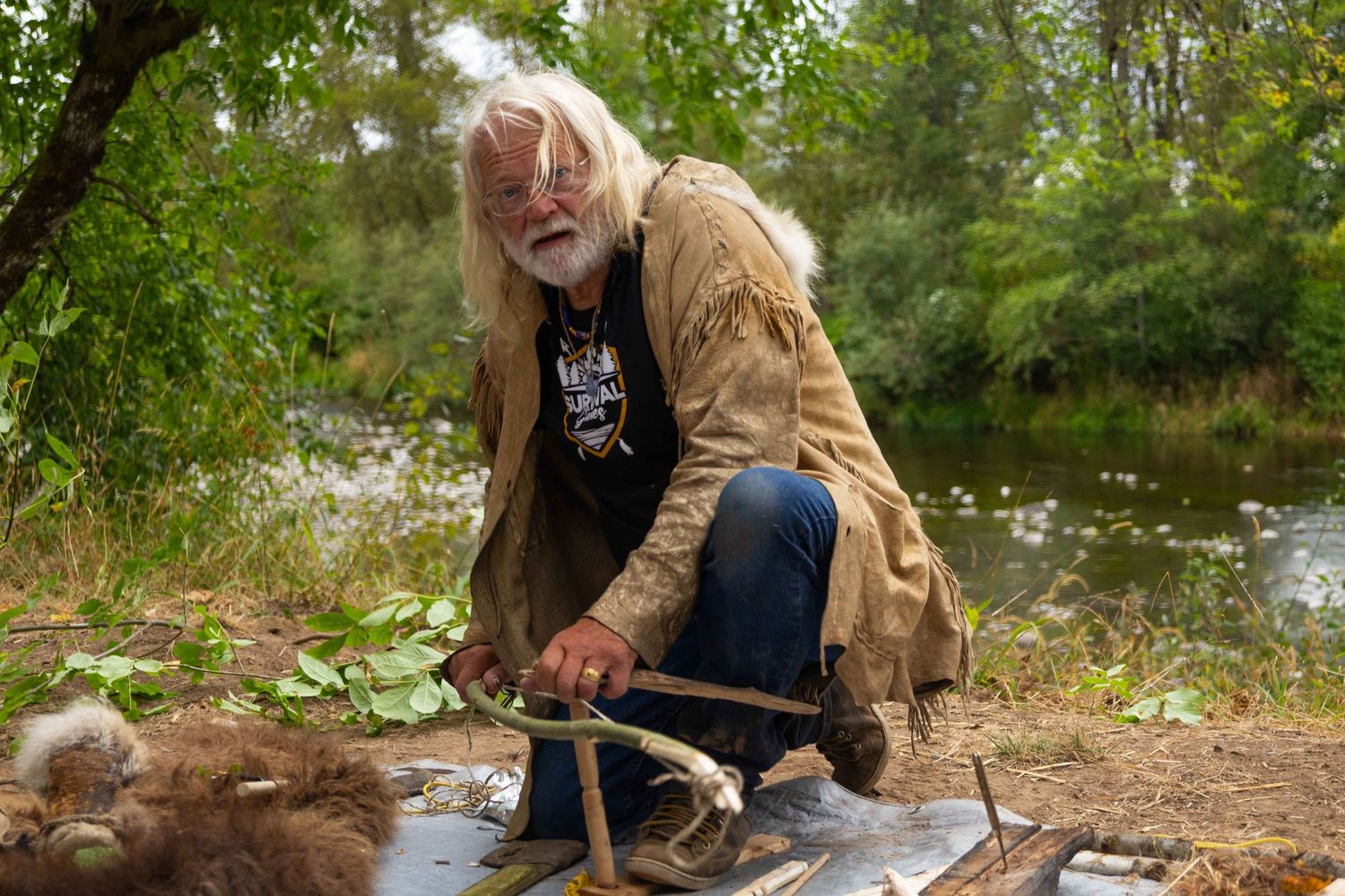 A person in a brown jacket and jeans crouches near a stream while fishing in a wooded area.