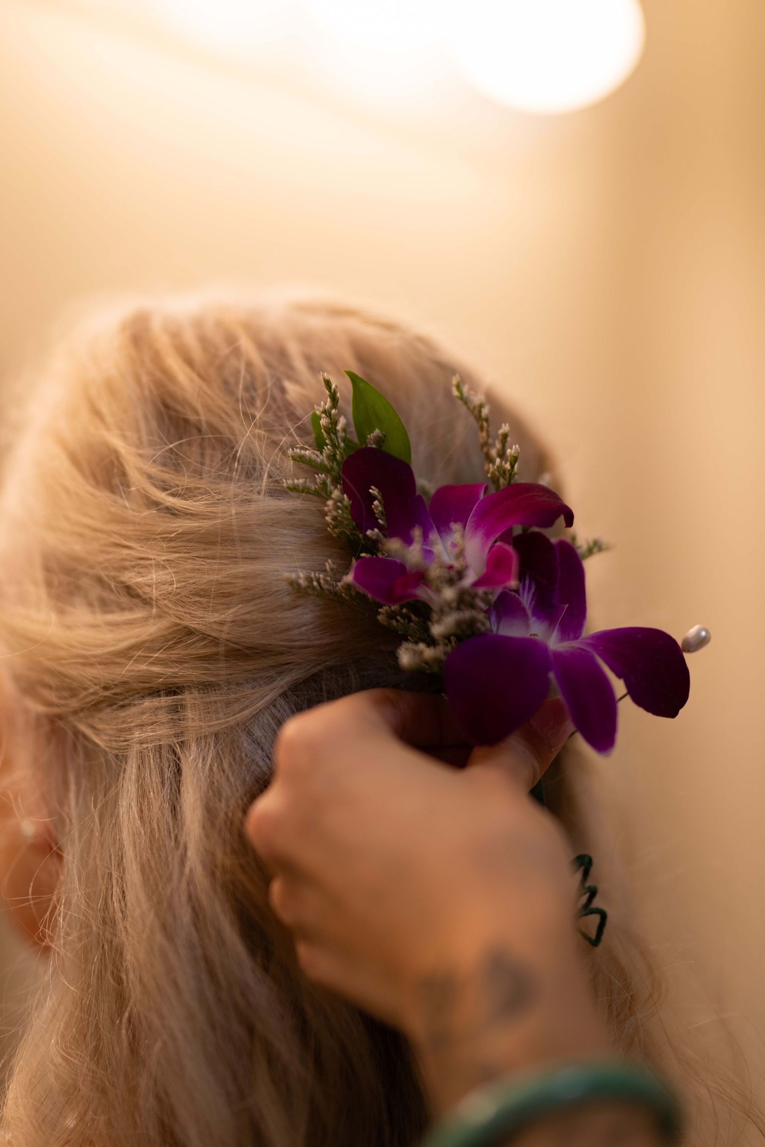 A hand places a purple orchid flower and greenery as a decorative hair accessory in blonde hair.