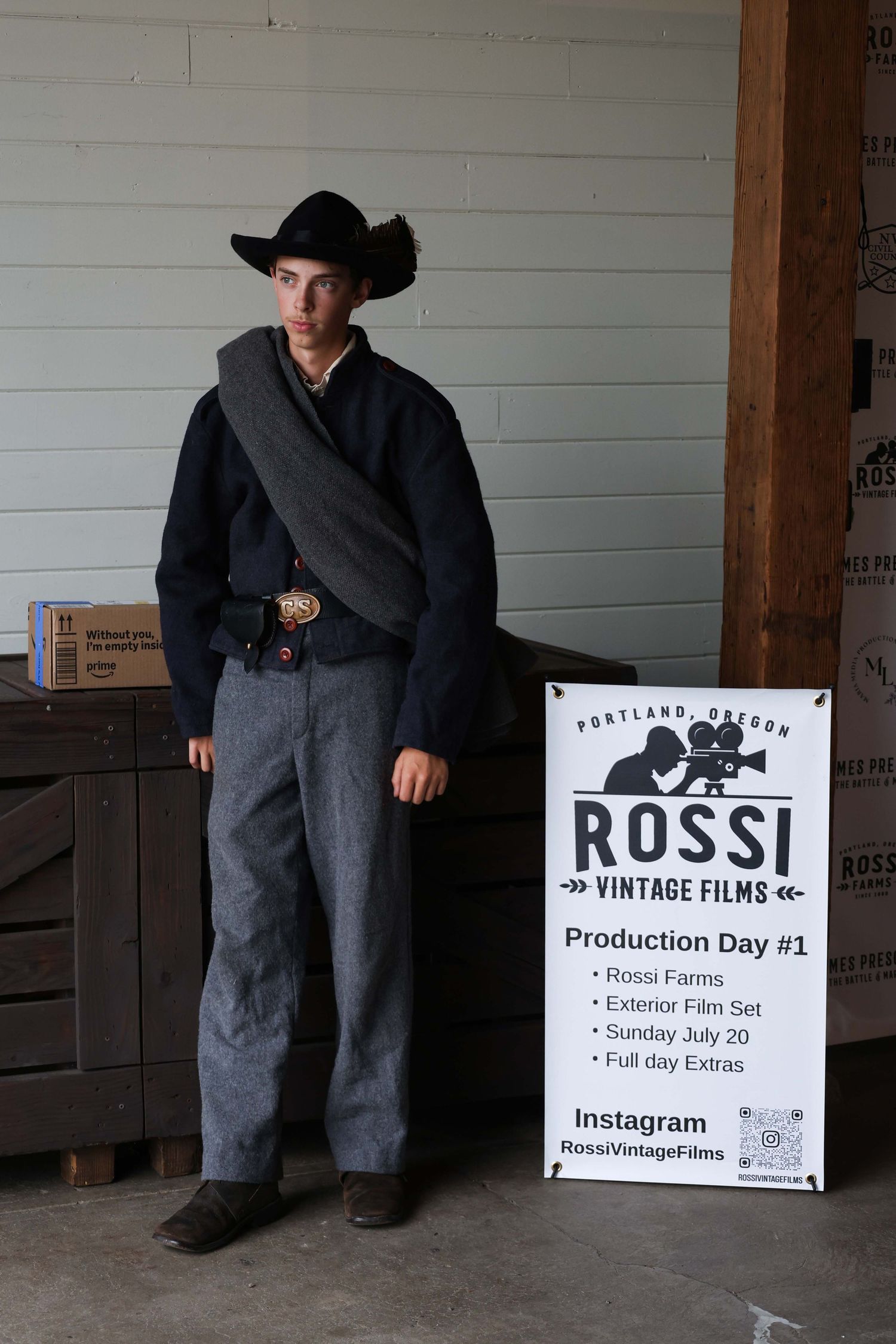 A person in period costume poses next to a Rossi Vintage Films production sign against a white wooden wall.