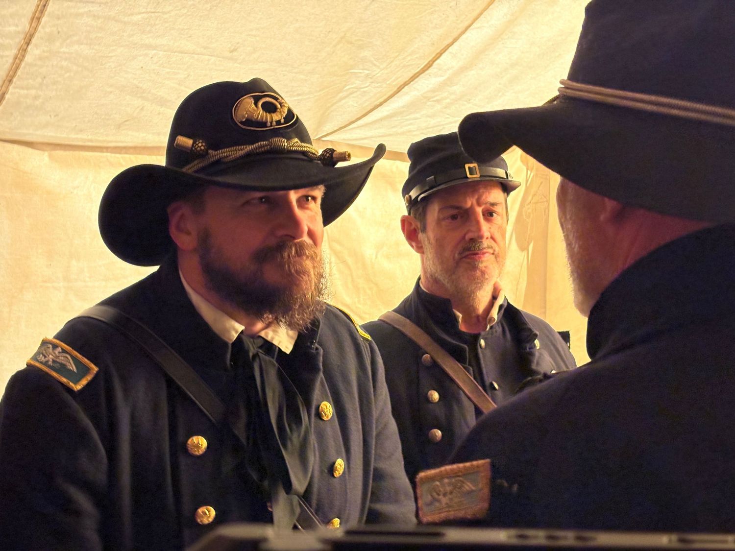 Civil War reenactors in Union military uniforms and period hats gather inside a canvas tent during a historical event.