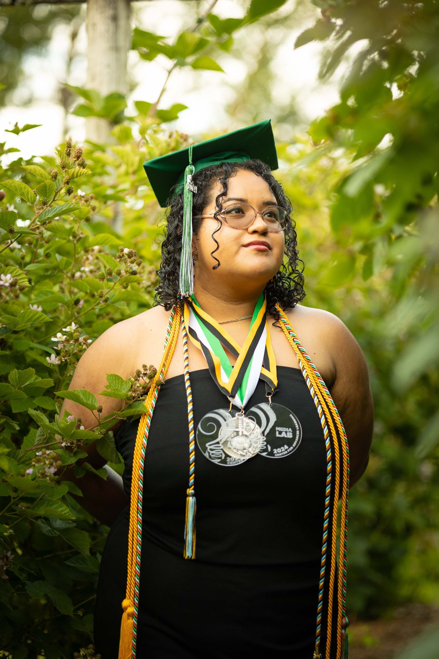 Graduate wearing academic regalia with medals and cords stands among green foliage in outdoor portrait.
