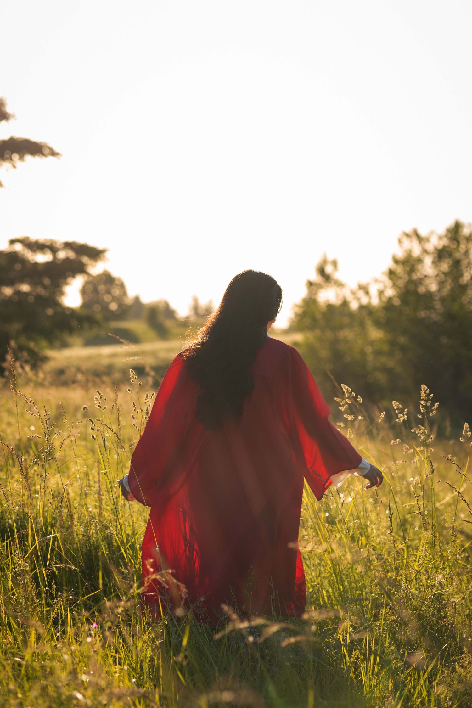 Person in flowing red dress stands in golden sunlit meadow during sunset with tall grass and trees in background.