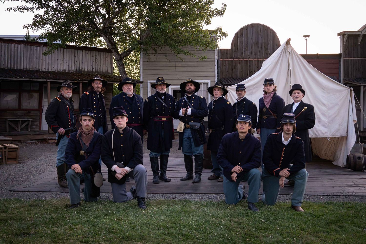 Group of men in dark uniforms stand together in front of white canvas tents at an outdoor event.