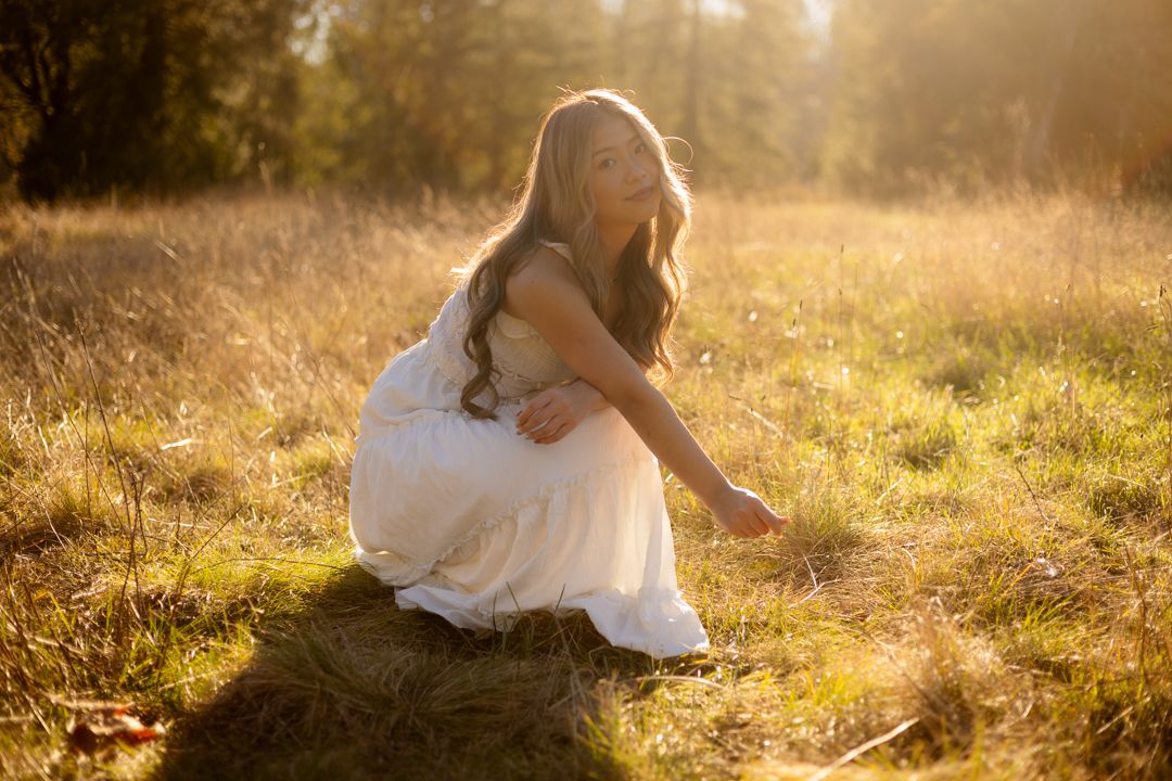 Woman in white dress sitting in golden sunlit field, long hair flowing, serene outdoor portrait photography.