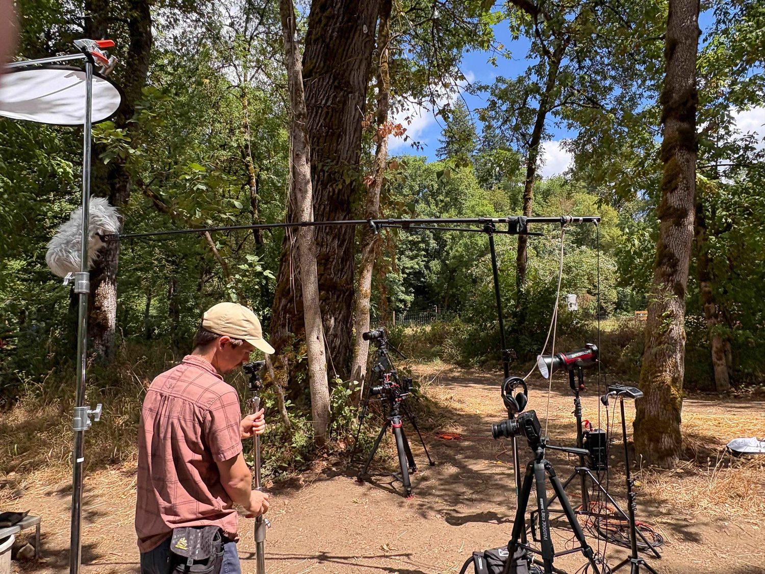 Outdoor film production setup in a wooded area with lighting equipment and camera gear arranged between trees.