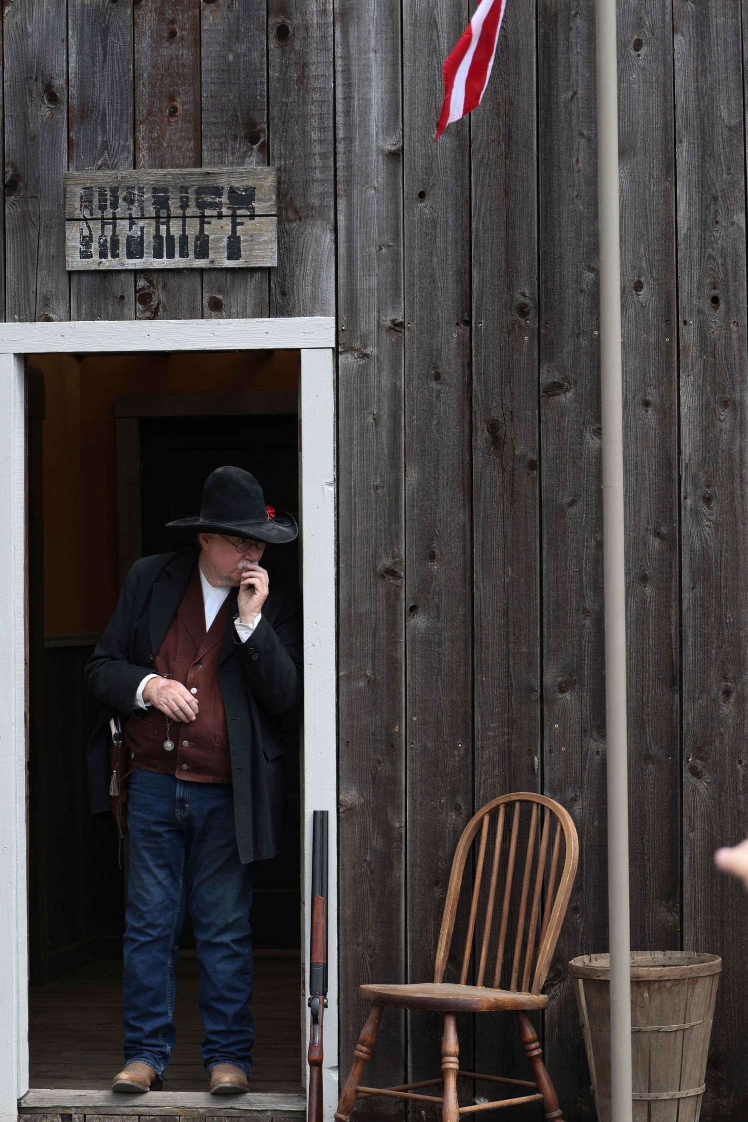 A figure in western attire stands in a rustic wooden doorway with a chair and American flag visible outside.