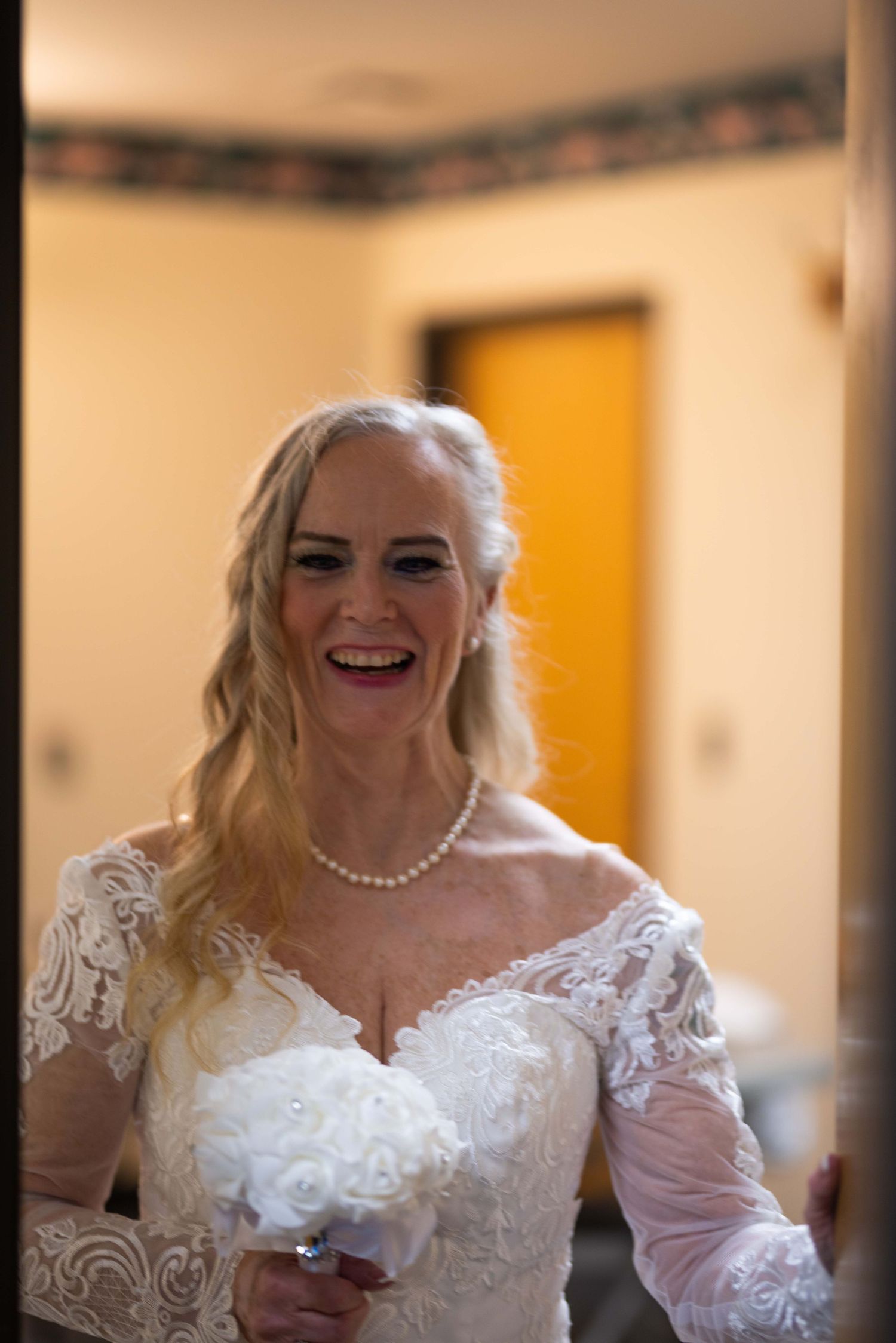 A bride wearing a white lace wedding dress with sheer sleeves and pearl necklace smiles joyfully.