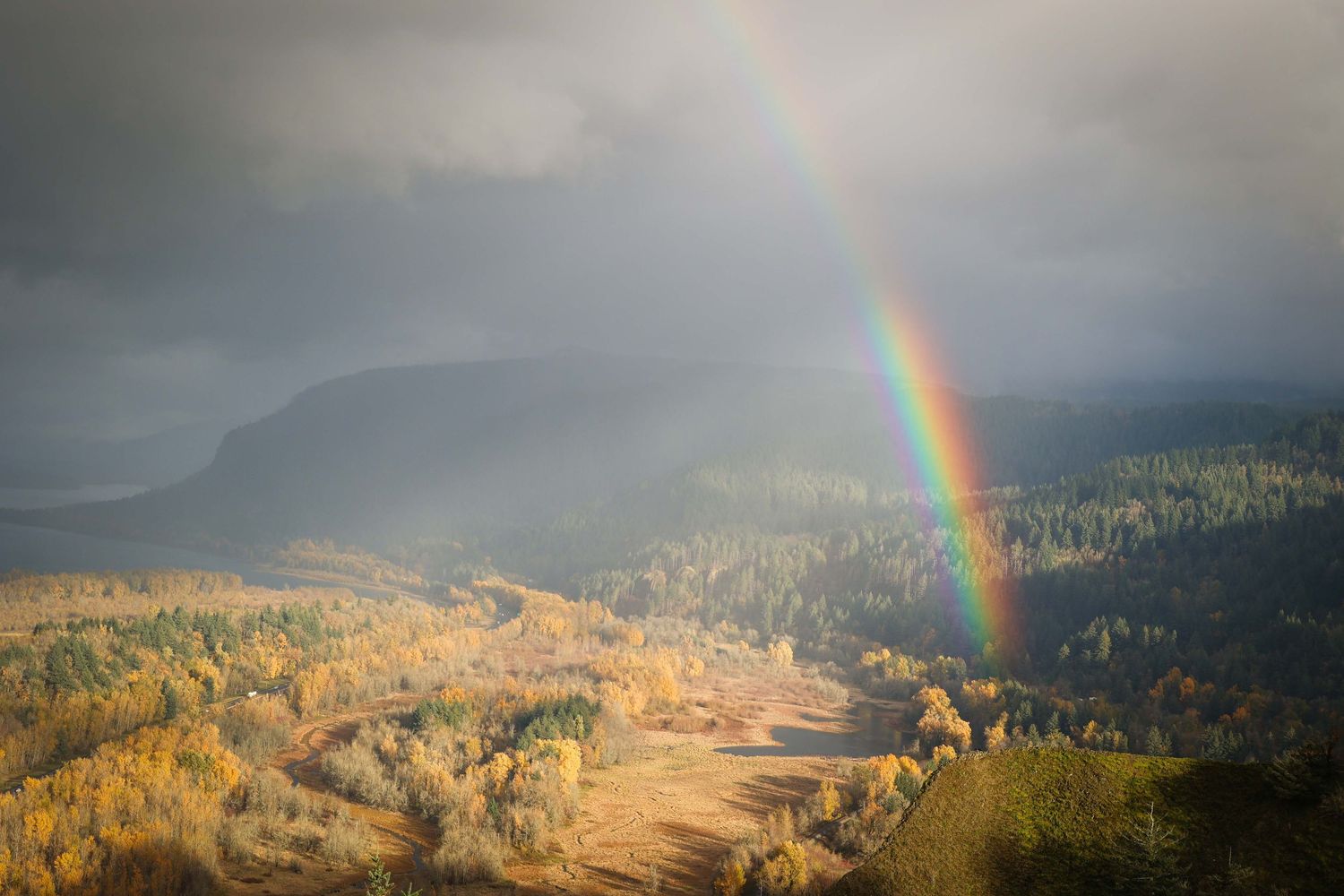 Vibrant rainbow arcs across a misty mountain valley with autumn foliage and sunlit slopes.