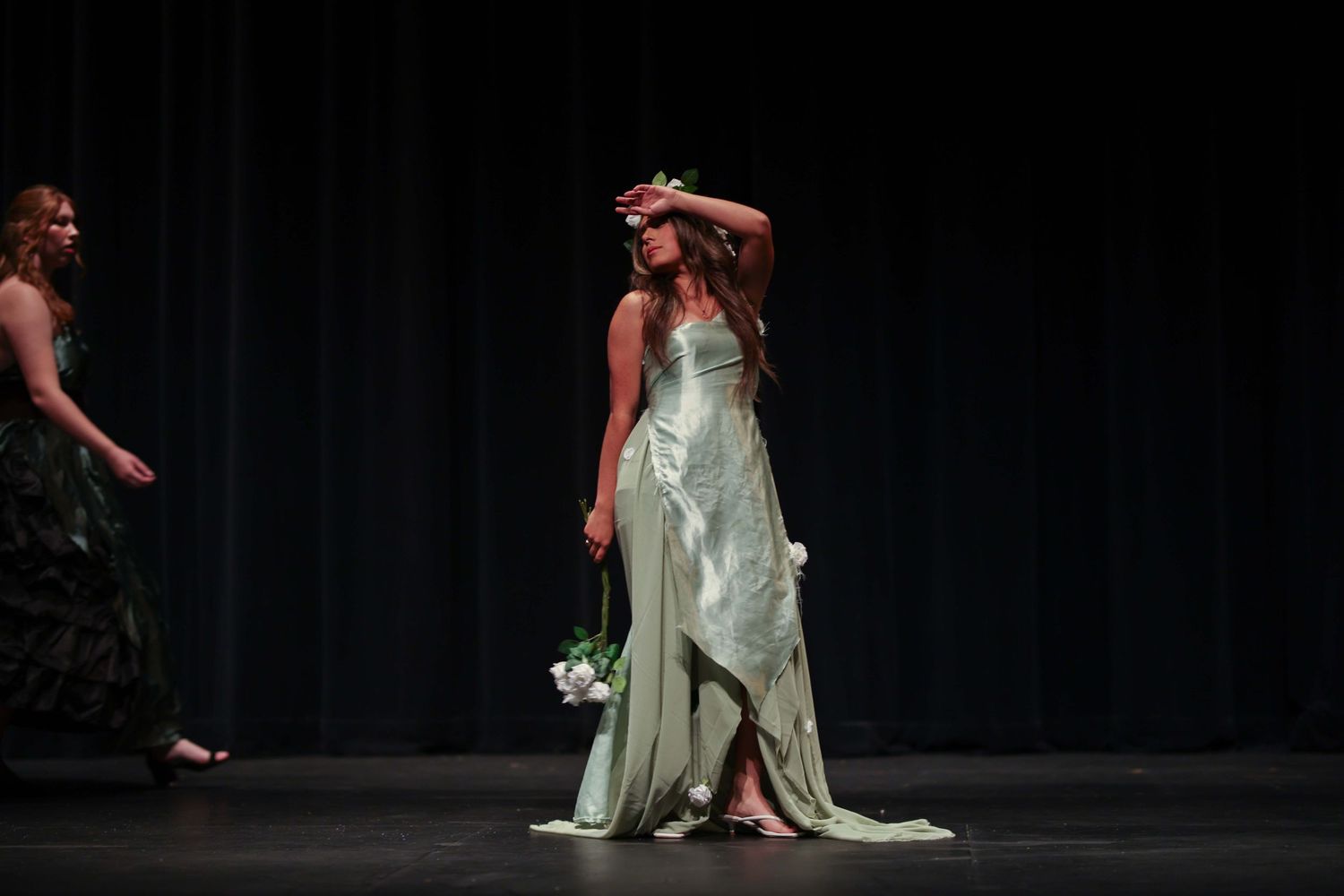 A ballet dancer in a flowing mint green dress performs graceful movements on a dark stage during a performance.