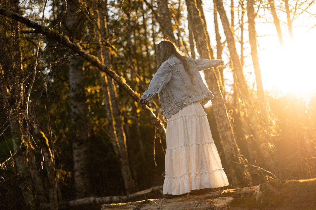 Woman in white skirt and denim jacket standing on log in sunlit forest at golden hour.