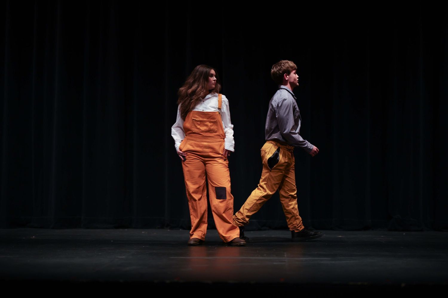 Two performers in orange and brown outfits stand back-to-back on a dark stage during a theatrical performance.