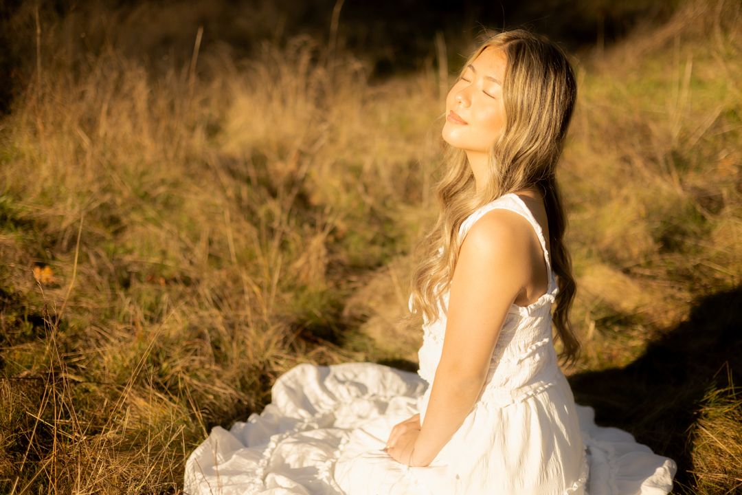 Young woman in white dress sits peacefully in golden sunlit field with eyes closed, long blonde hair flowing.