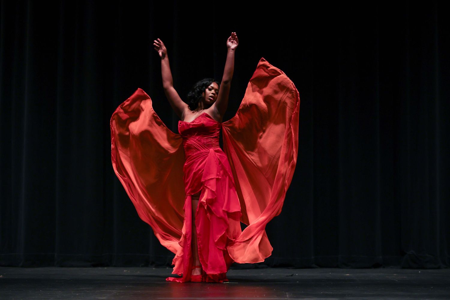 A dancer in flowing red fabric performs an expressive contemporary dance movement against a dark background.