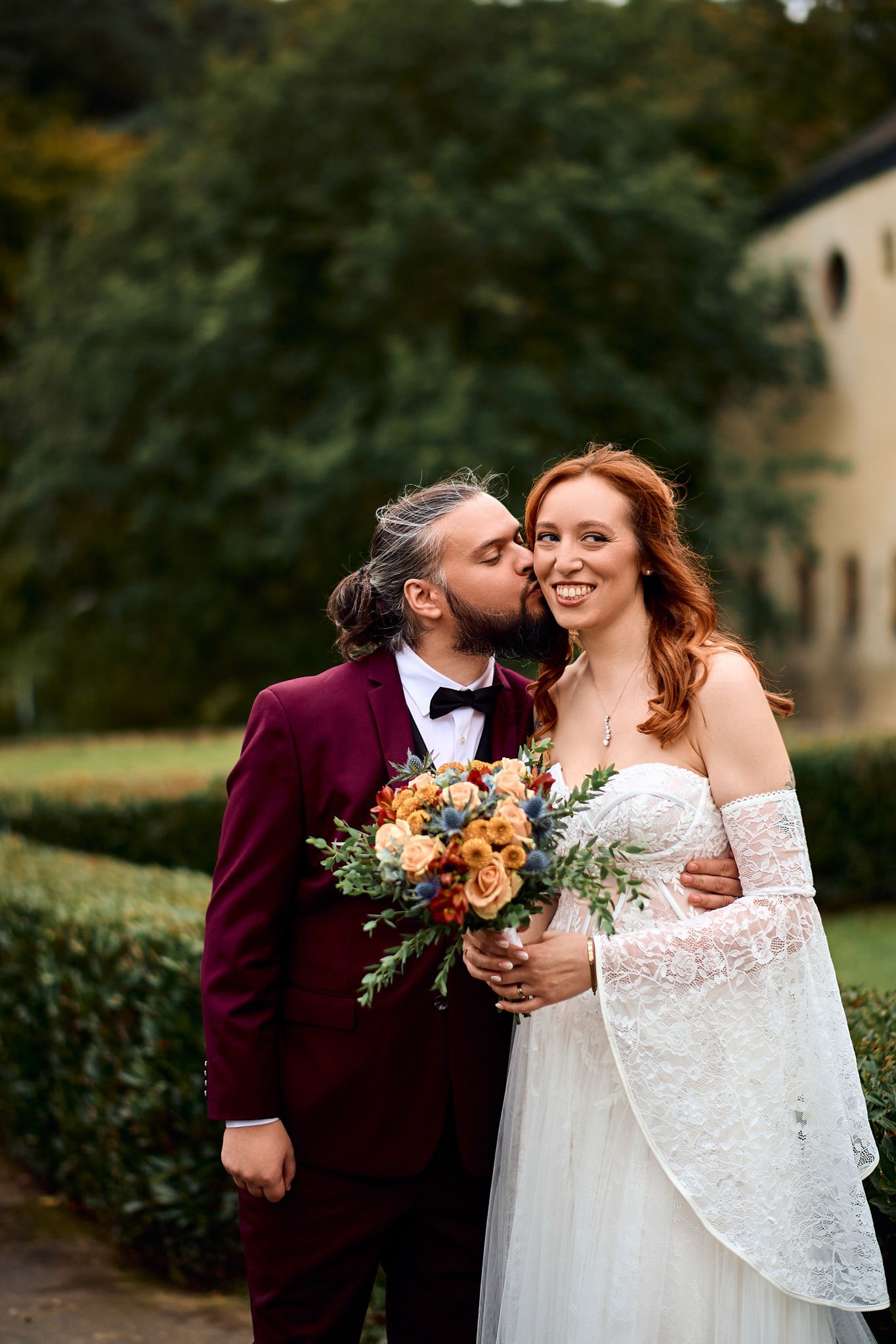 Romantic wedding couple embraces in a garden setting with burgundy and white attire.