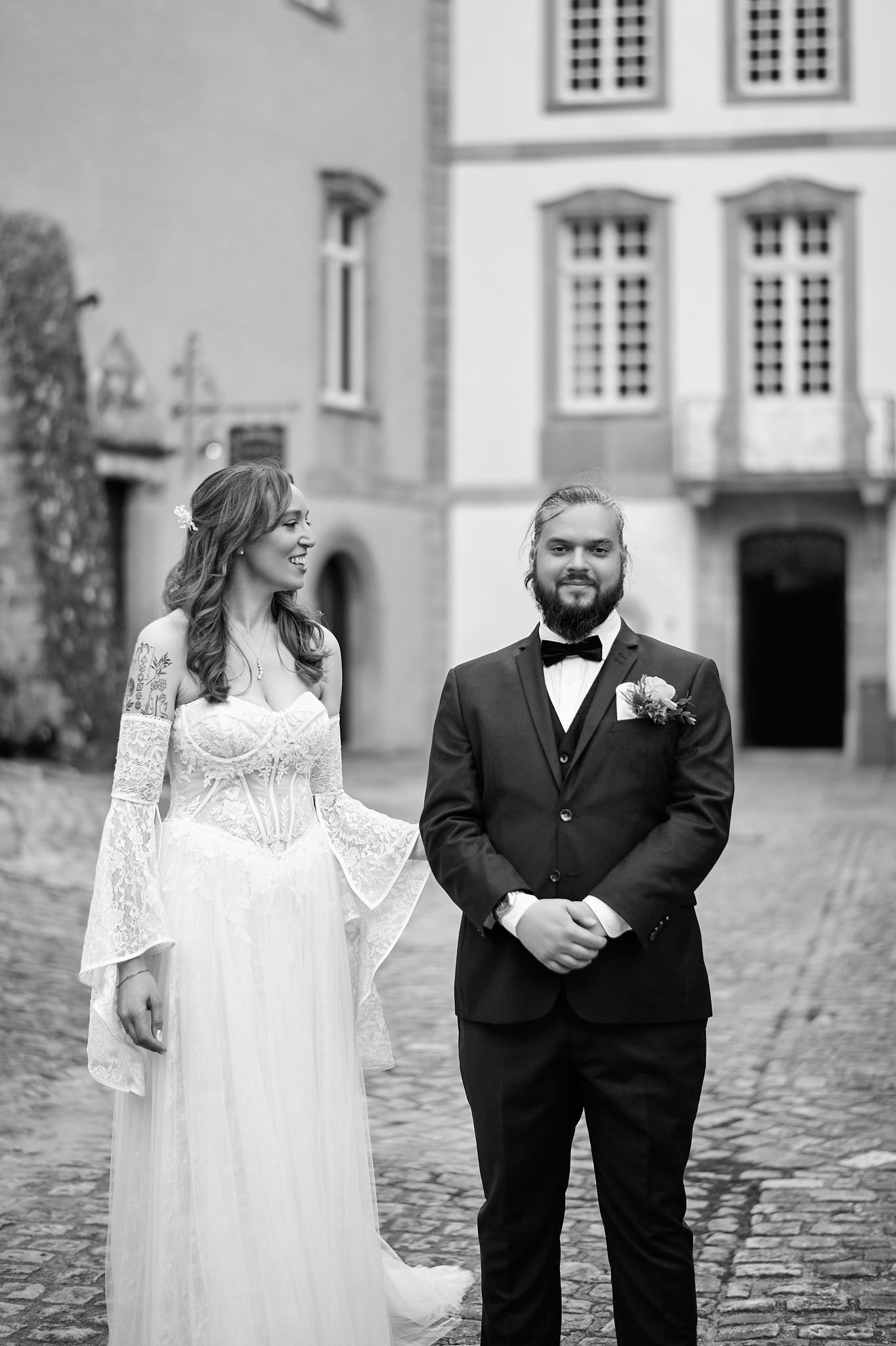 Wedding couple poses on cobblestone path outside historic building in formal attire.