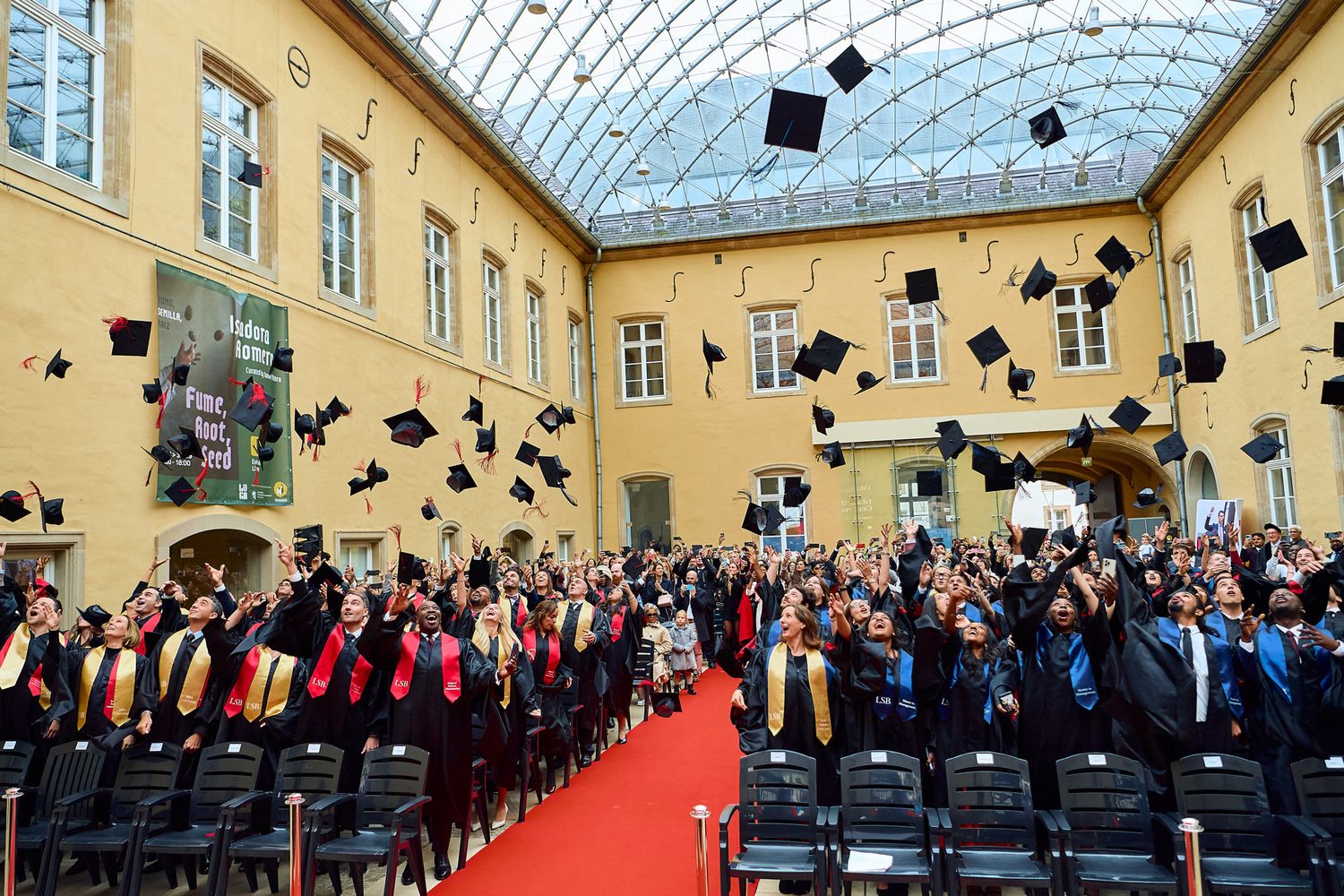 Graduates in black caps and gowns stand in rows during an indoor graduation ceremony under a glass ceiling.