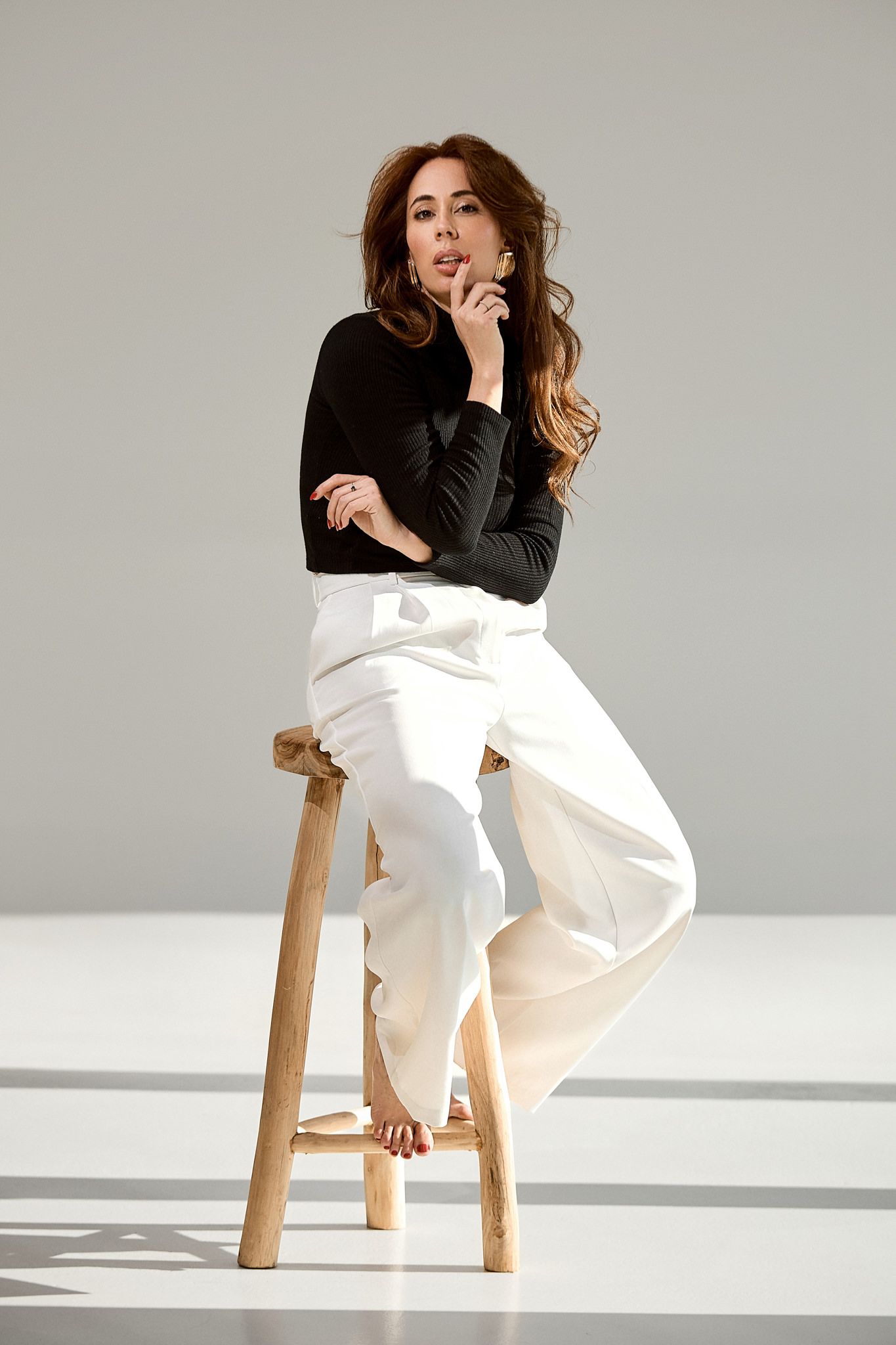 Fashion model poses on wooden stool wearing black top and flowing white pants in minimalist studio setting.