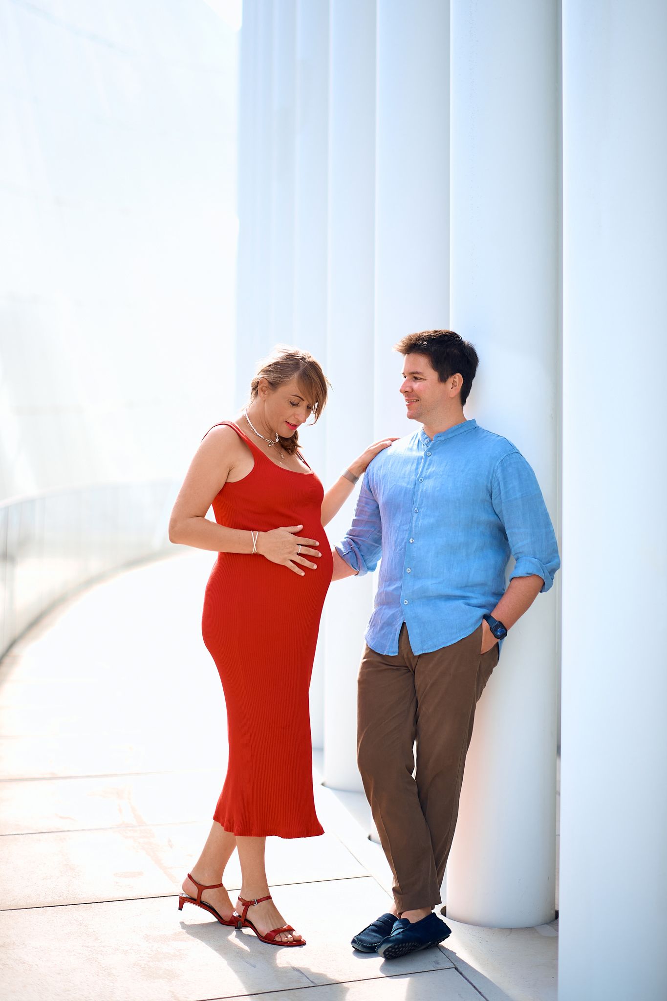 Couple in red dress and blue shirt posing playfully against white columns.