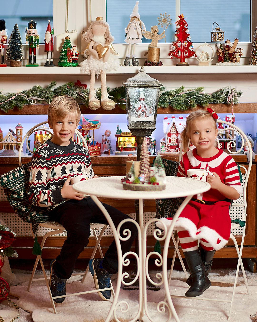 Two children in festive attire sit at a white table surrounded by Christmas decorations and holiday figurines.