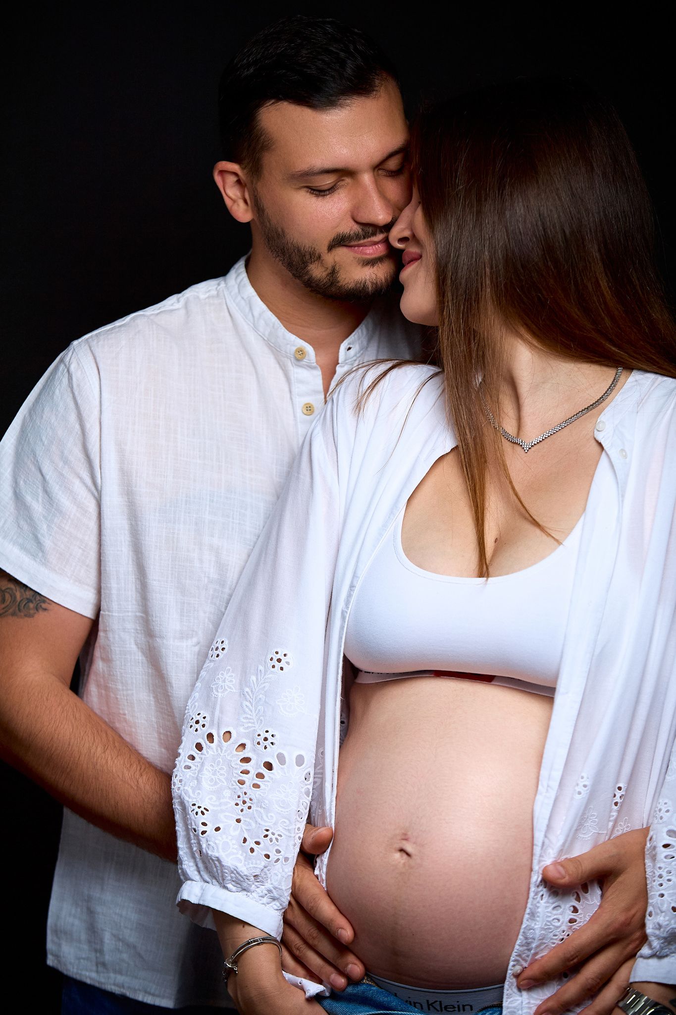 Intimate maternity photo session with couple in white clothing against dark background.
