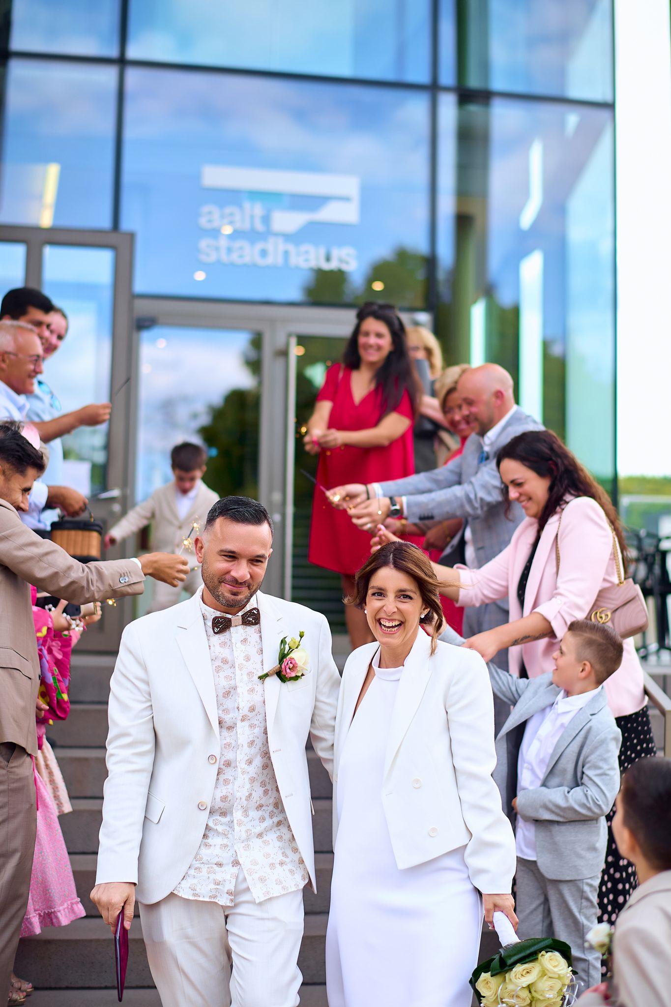 Wedding celebration outside modern glass building with guests in white and light colored attire.