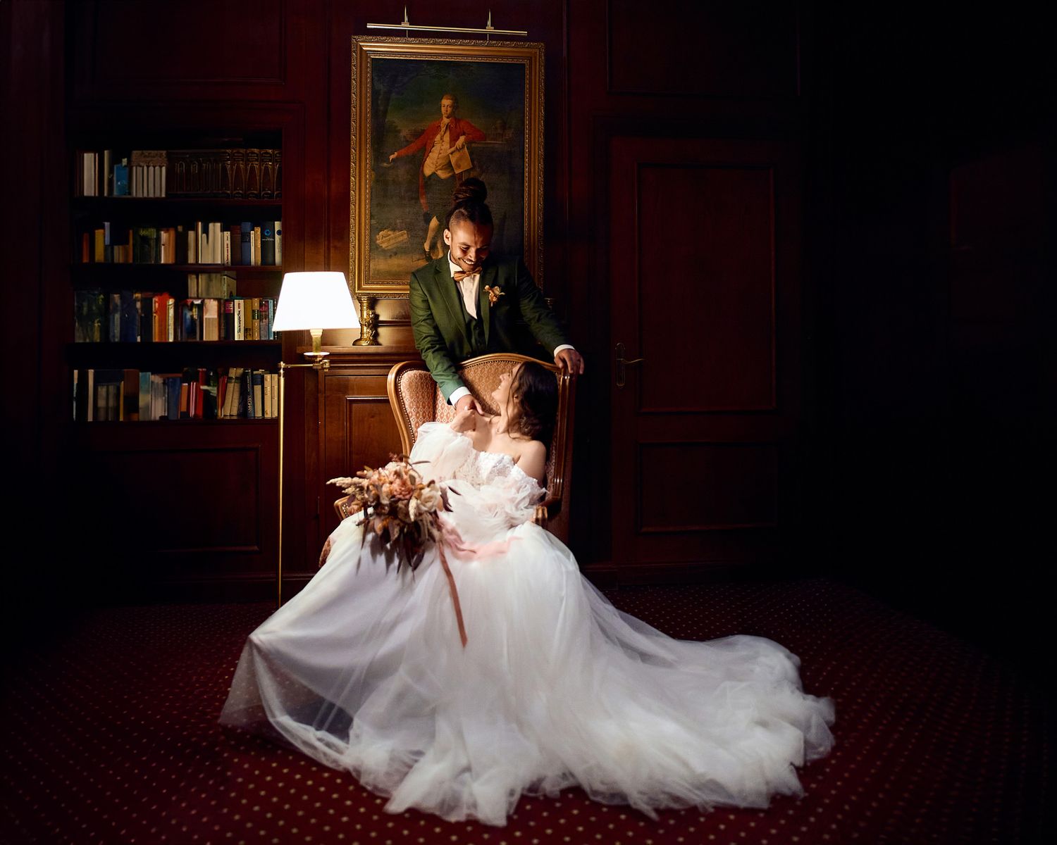 Dramatic wedding portrait in library with couple posed near antique chair and bookshelf.