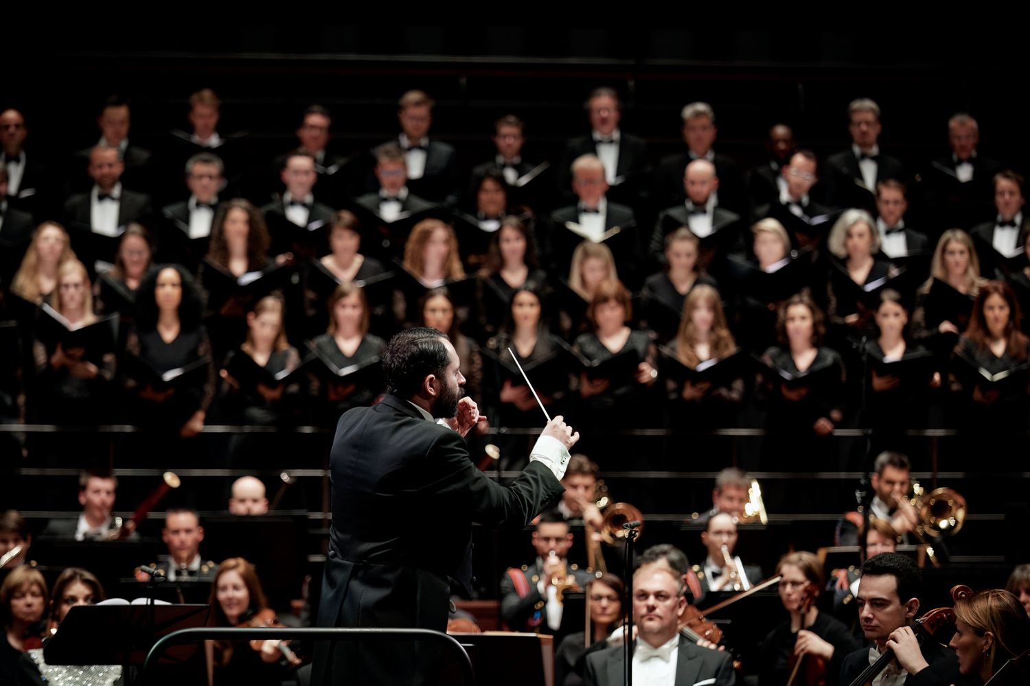 Orchestra and choir performers in formal black attire perform on stage in concert hall.