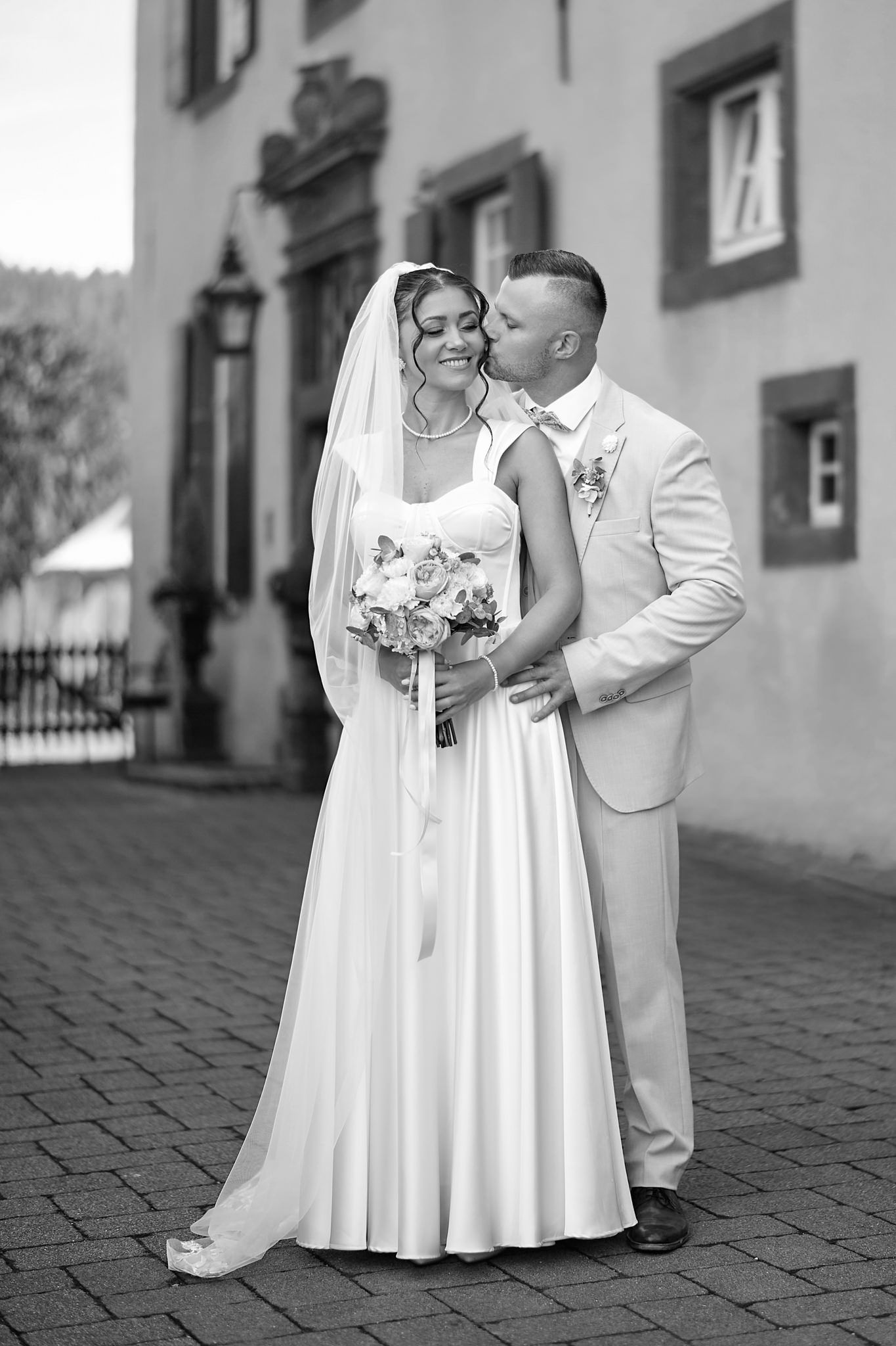 A couple in white wedding attire poses together on a cobblestone street.